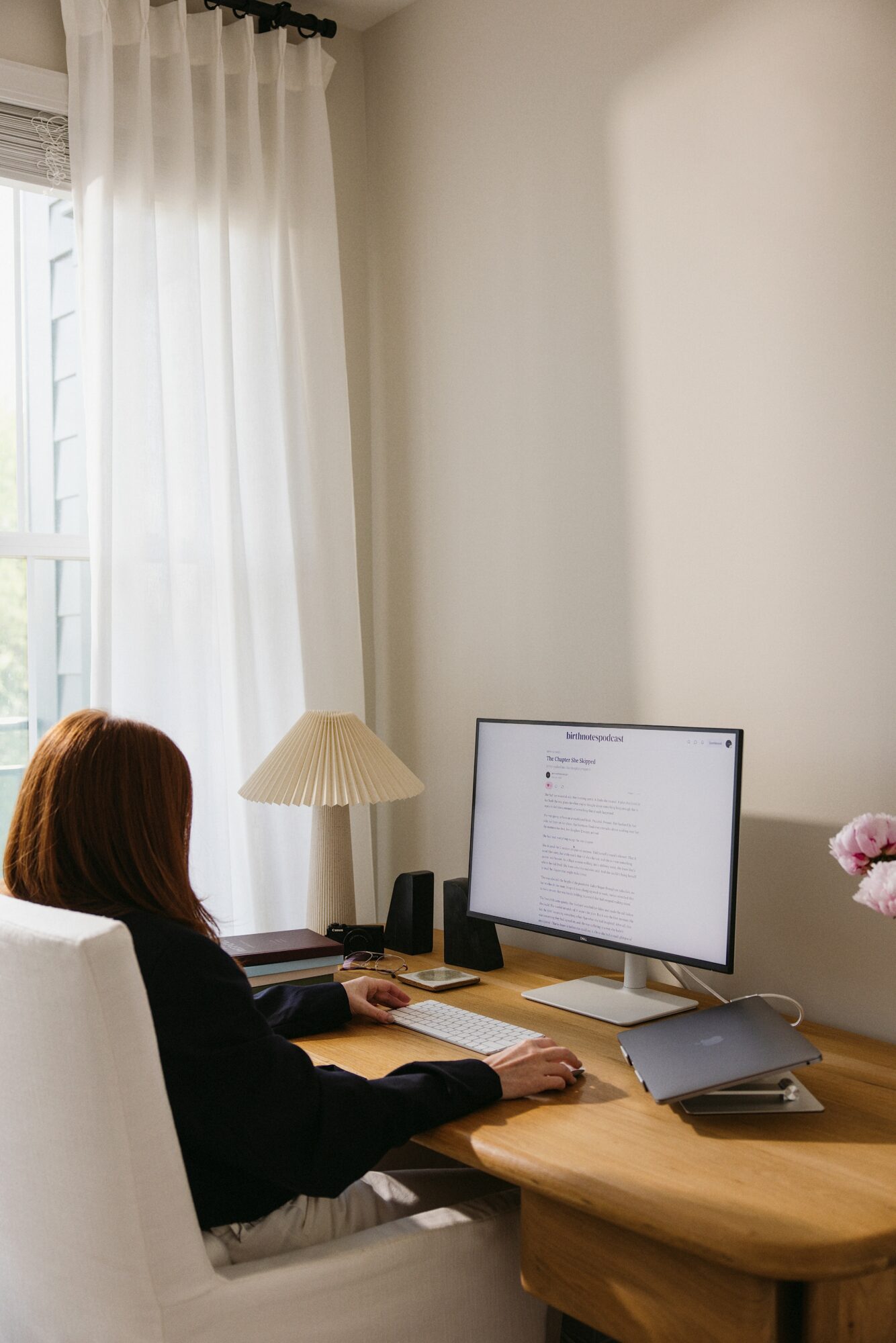 Person with red hair working at a desk with a computer monitor, lamp, and pink flowers, near a window with white curtains