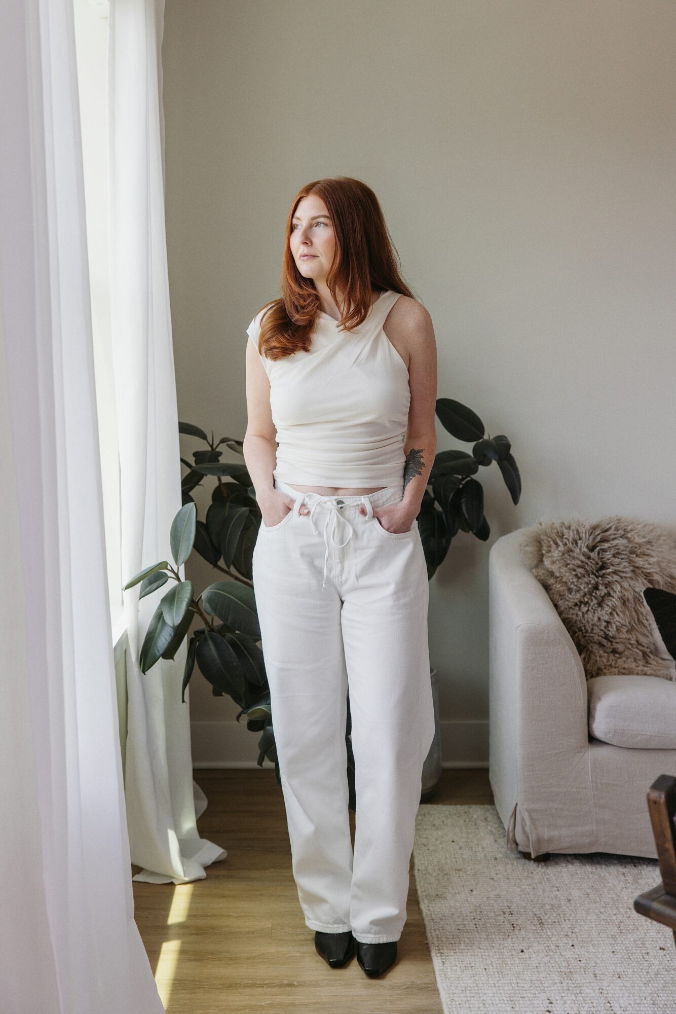 Woman with long red hair in white sleeveless top and white pants standing indoors near a window with plants and a sofa.