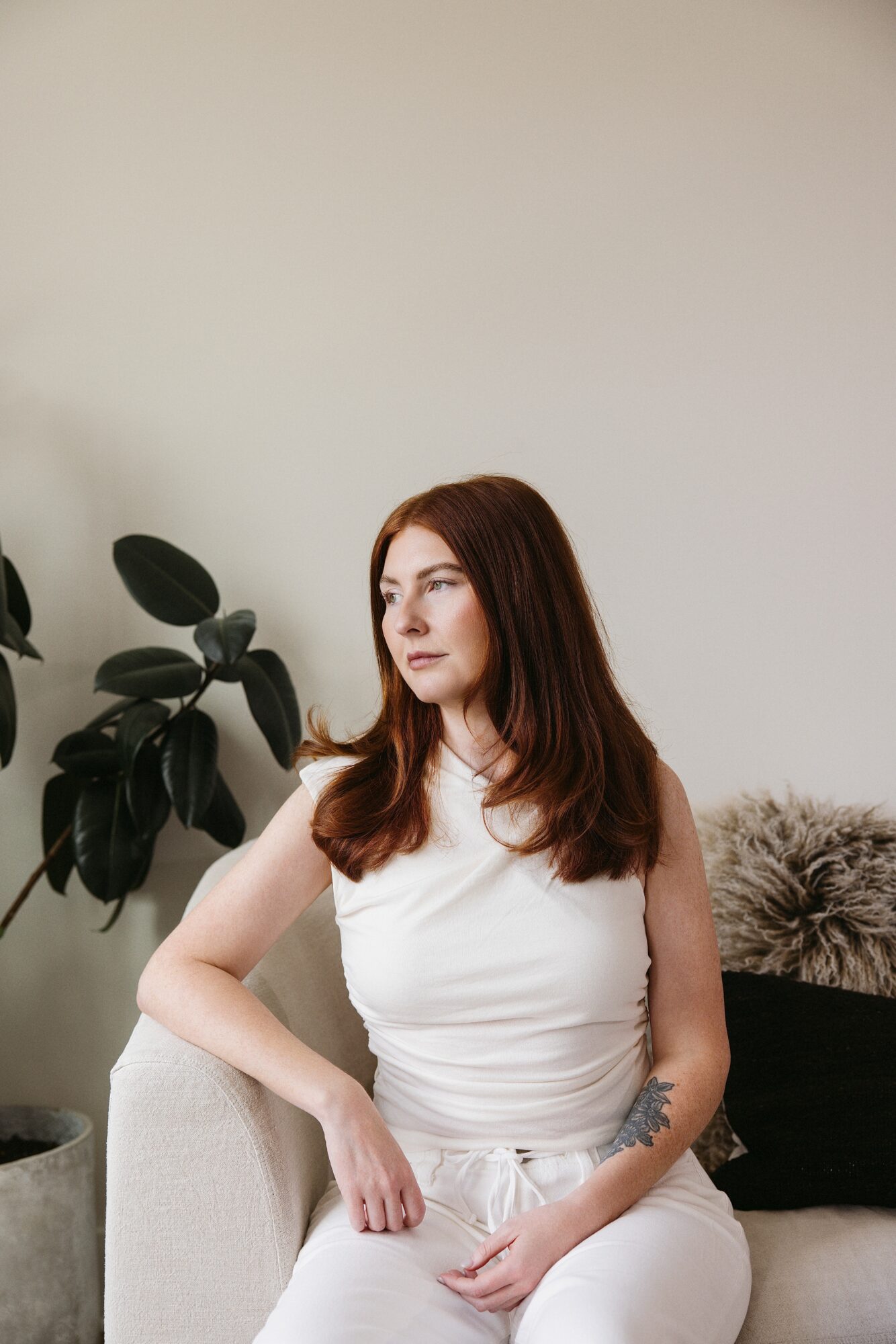 Woman with long red hair sitting on a light-colored sofa, wearing a white sleeveless top and pants, with a plant in the background.