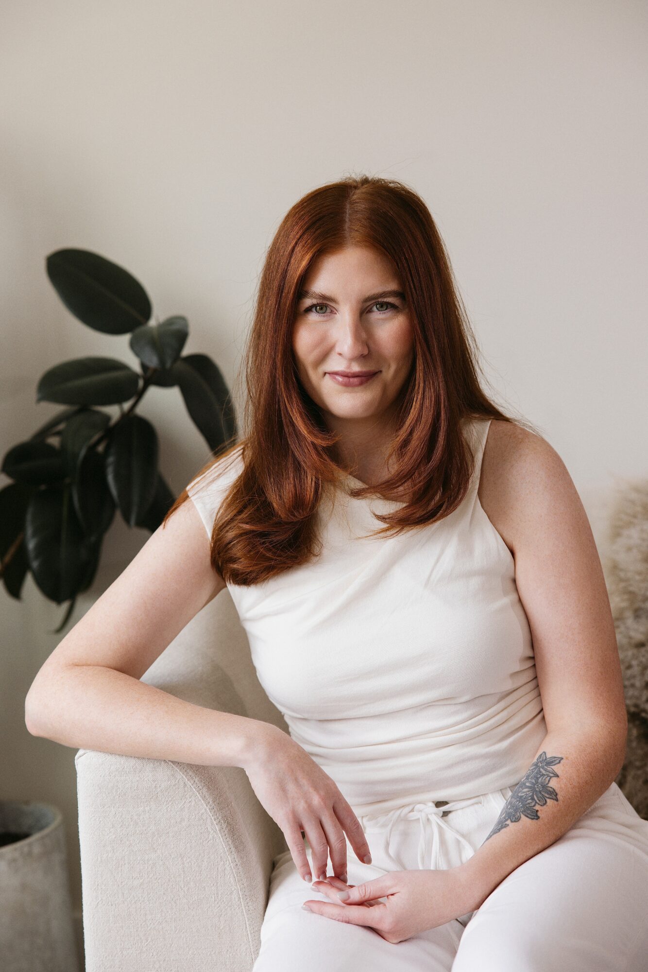 Woman with long red hair sitting on a light-colored sofa, smiling, with a plant in the background.