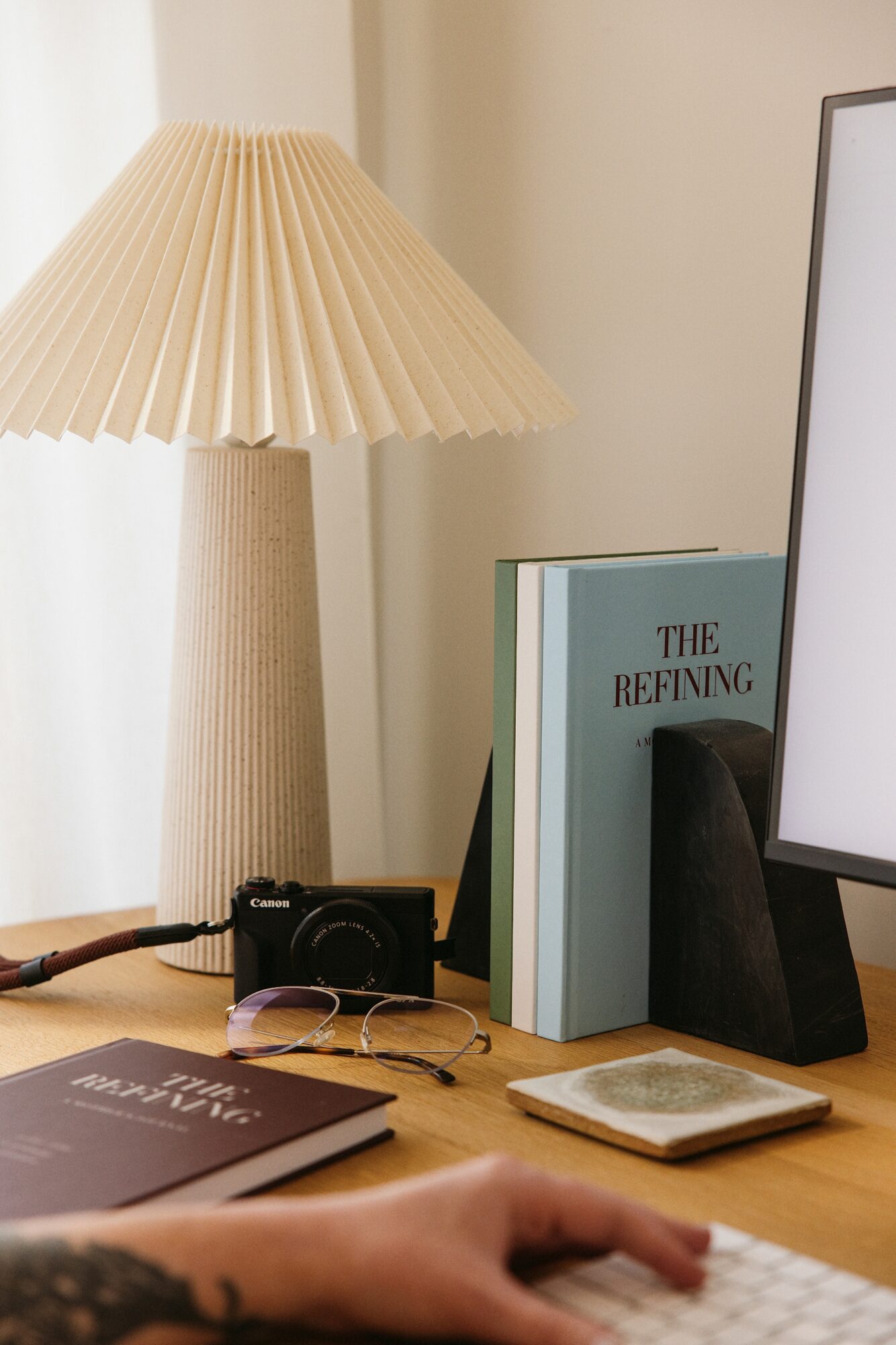 Desk with a lamp, a book titled 'The Refining,' a camera, glasses, and a hand on a keyboard.