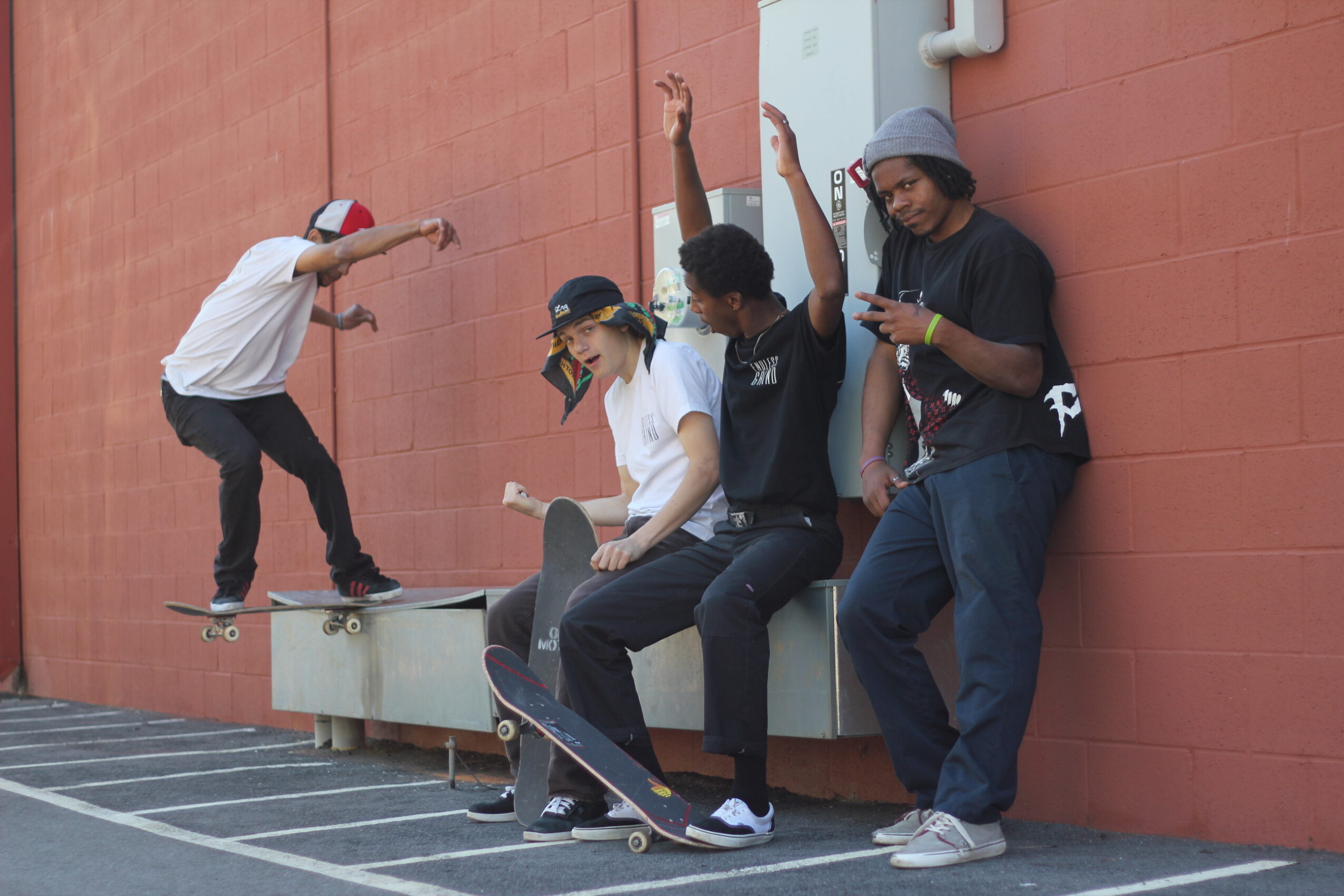 Four young people leaning against a red wall, one skateboarding, others using phones, with a skateboard on the ground.