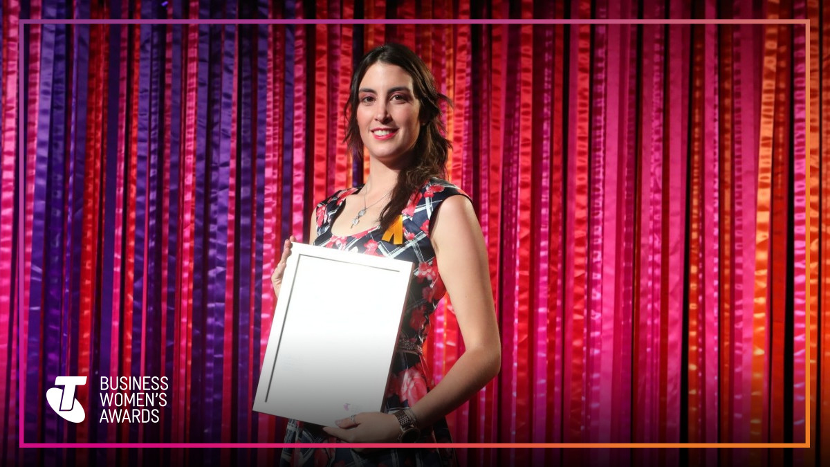 Woman holding an award plaque on stage with a colorful curtain background, smiling, at the Business Women's Awards.