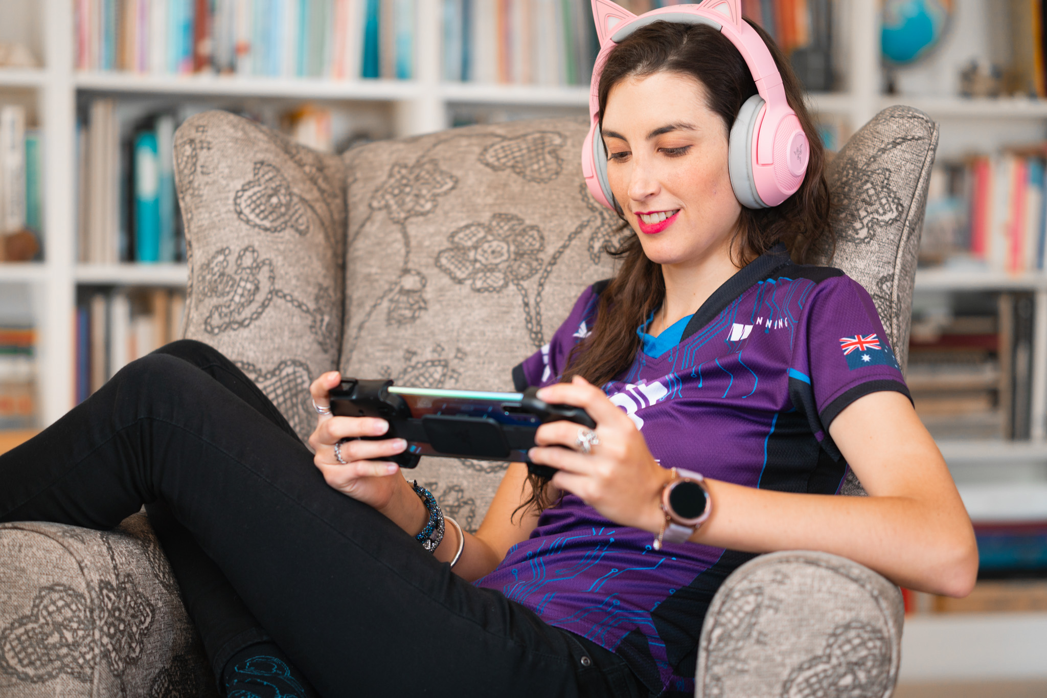 Young woman with headphones playing a handheld gaming console on a sofa in a library, smiling.