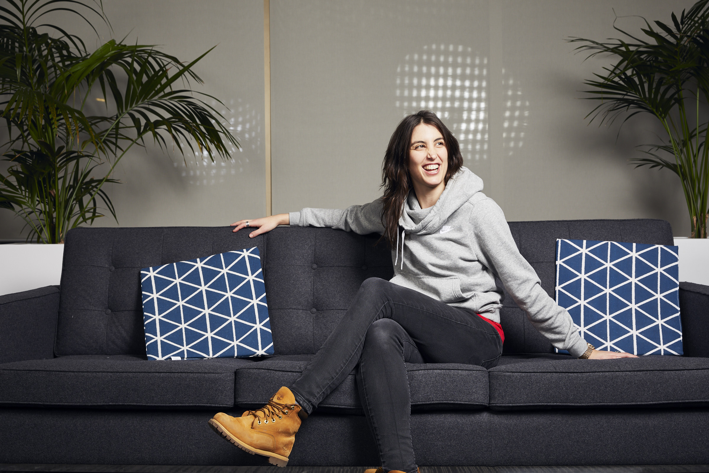 Woman sitting on a dark sofa with two blue patterned pillows, smiling, in a modern room with plants.