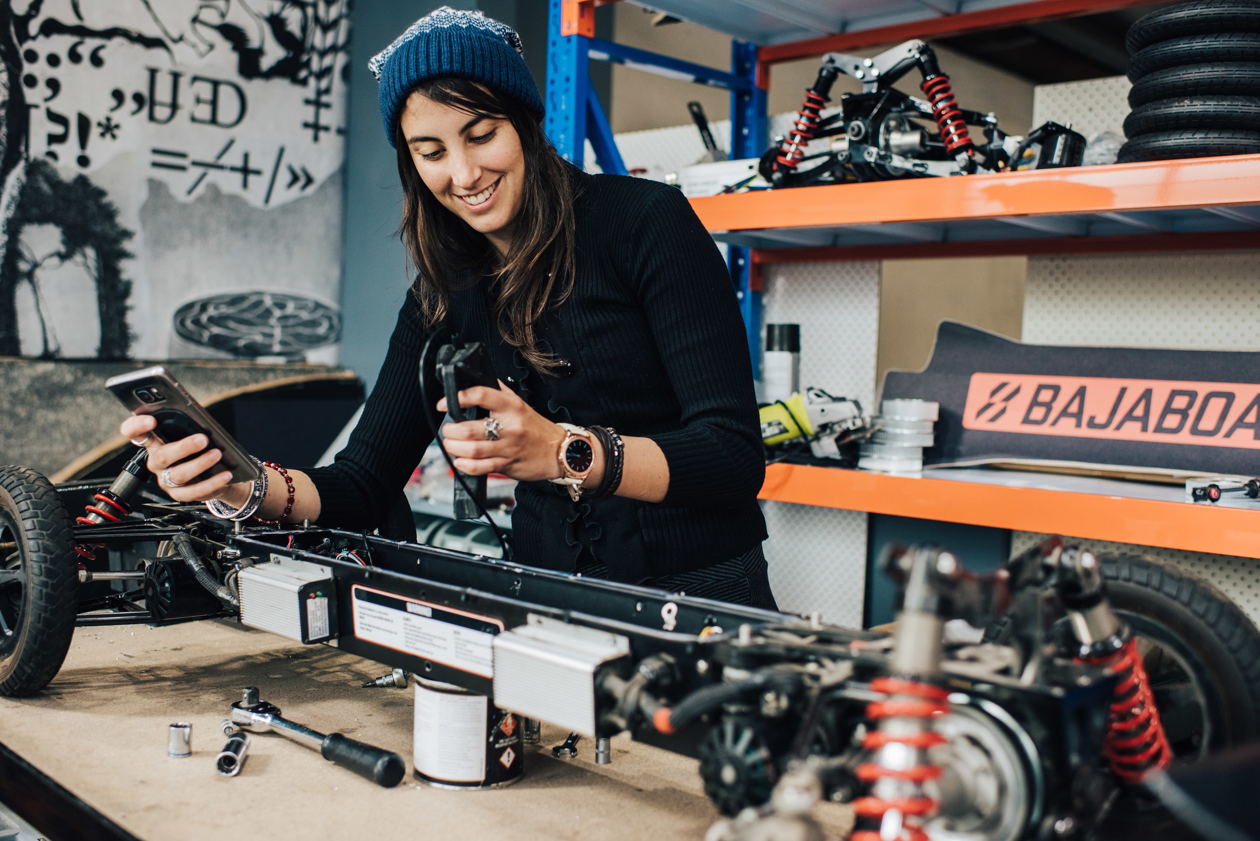 Woman in a blue beanie working on a bicycle frame in a workshop, holding a smartphone and a tool, smiling.