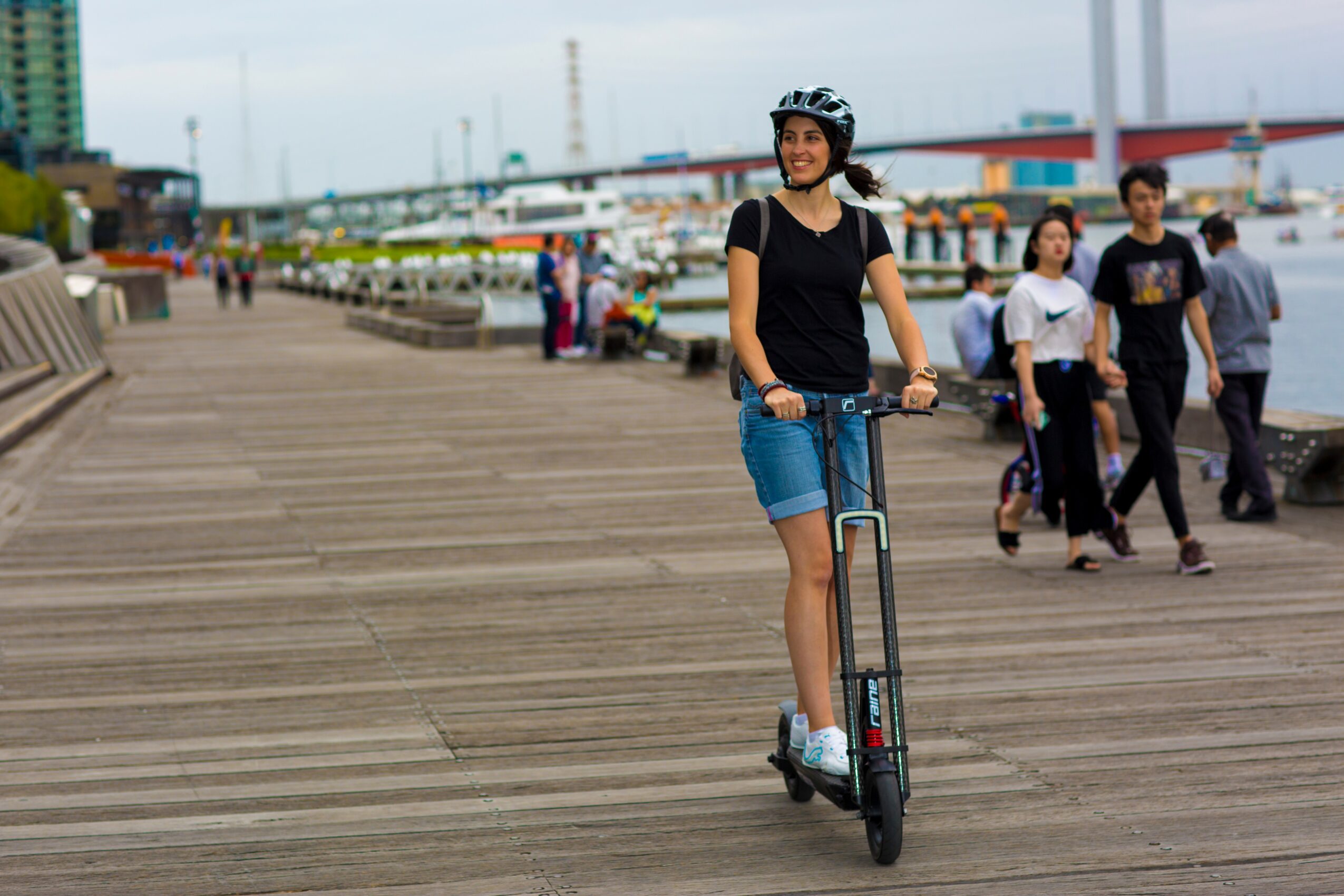 Woman riding a scooter on a wooden boardwalk, smiling, with people walking in the background, overcast sky.