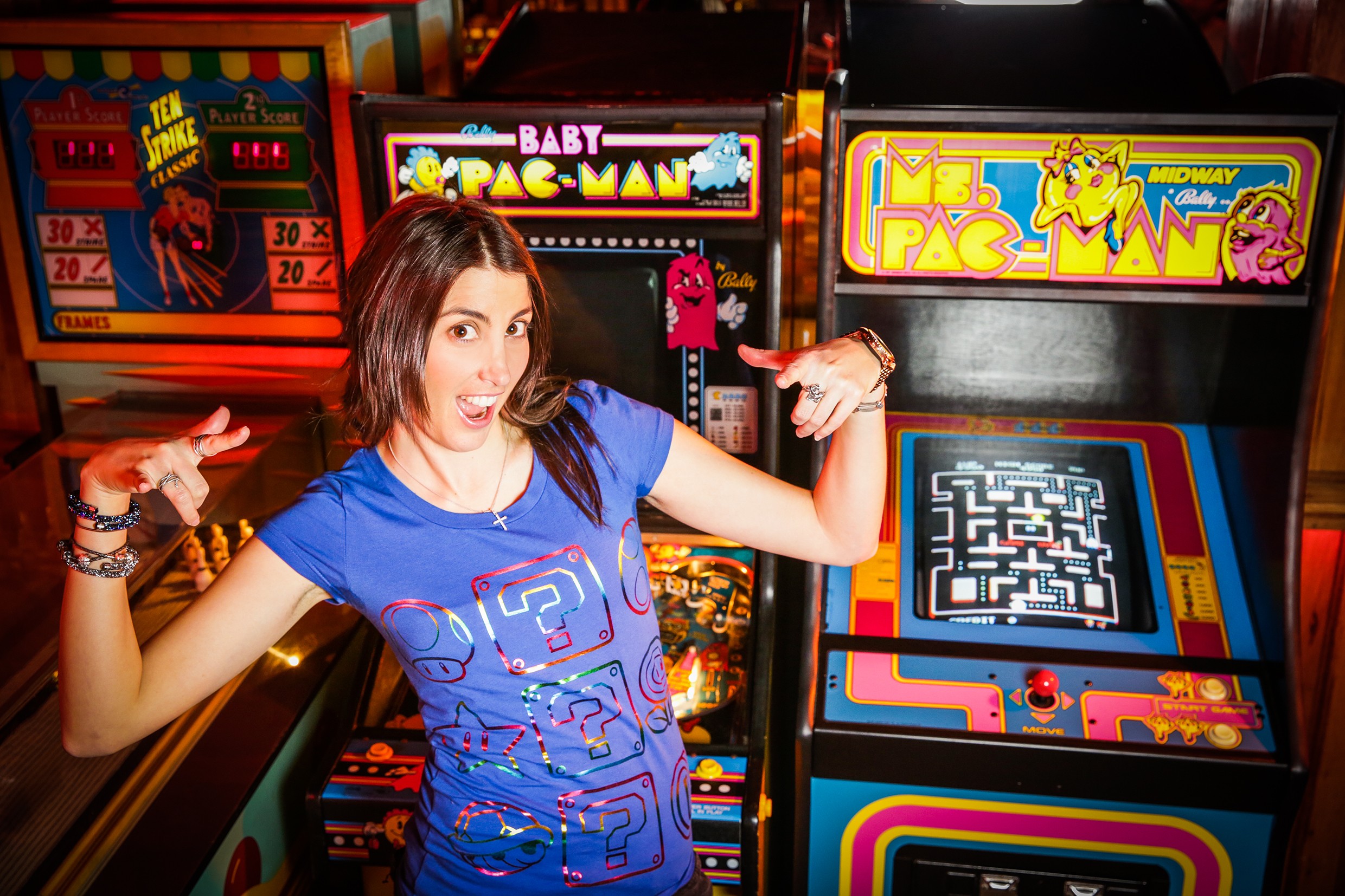 Woman in blue shirt pointing at arcade game with Pac-Man and Ms. Pac-Man signs behind her.