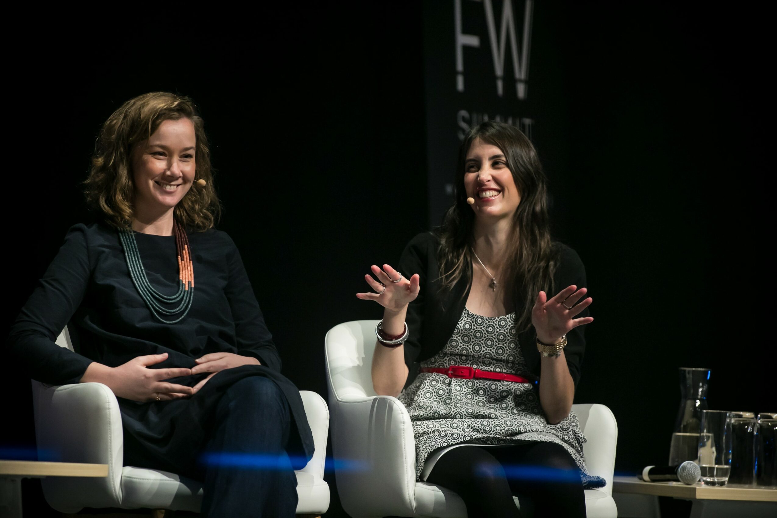 Two women sitting on white chairs, smiling and talking, with a dark background and a glass of water on a table nearby.
