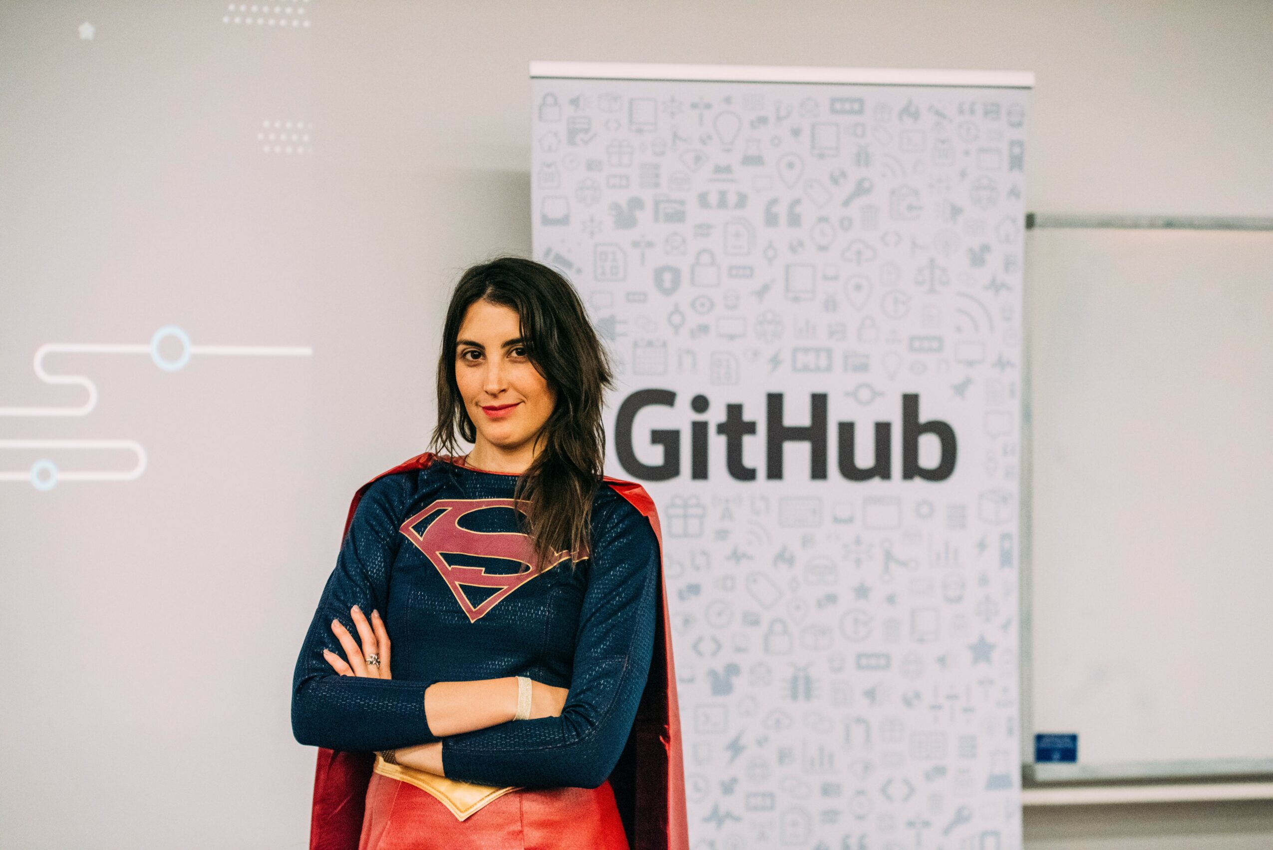 Woman with dark hair wearing a Superman logo shirt and dark jacket standing in front of a GitHub banner.