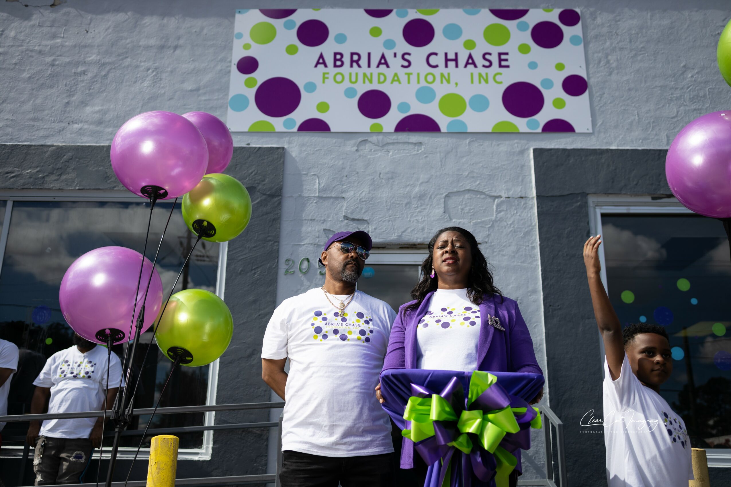 Two women and a man stand outside a building with balloons and a sign for Arbia's Chase Foundation, Inc.