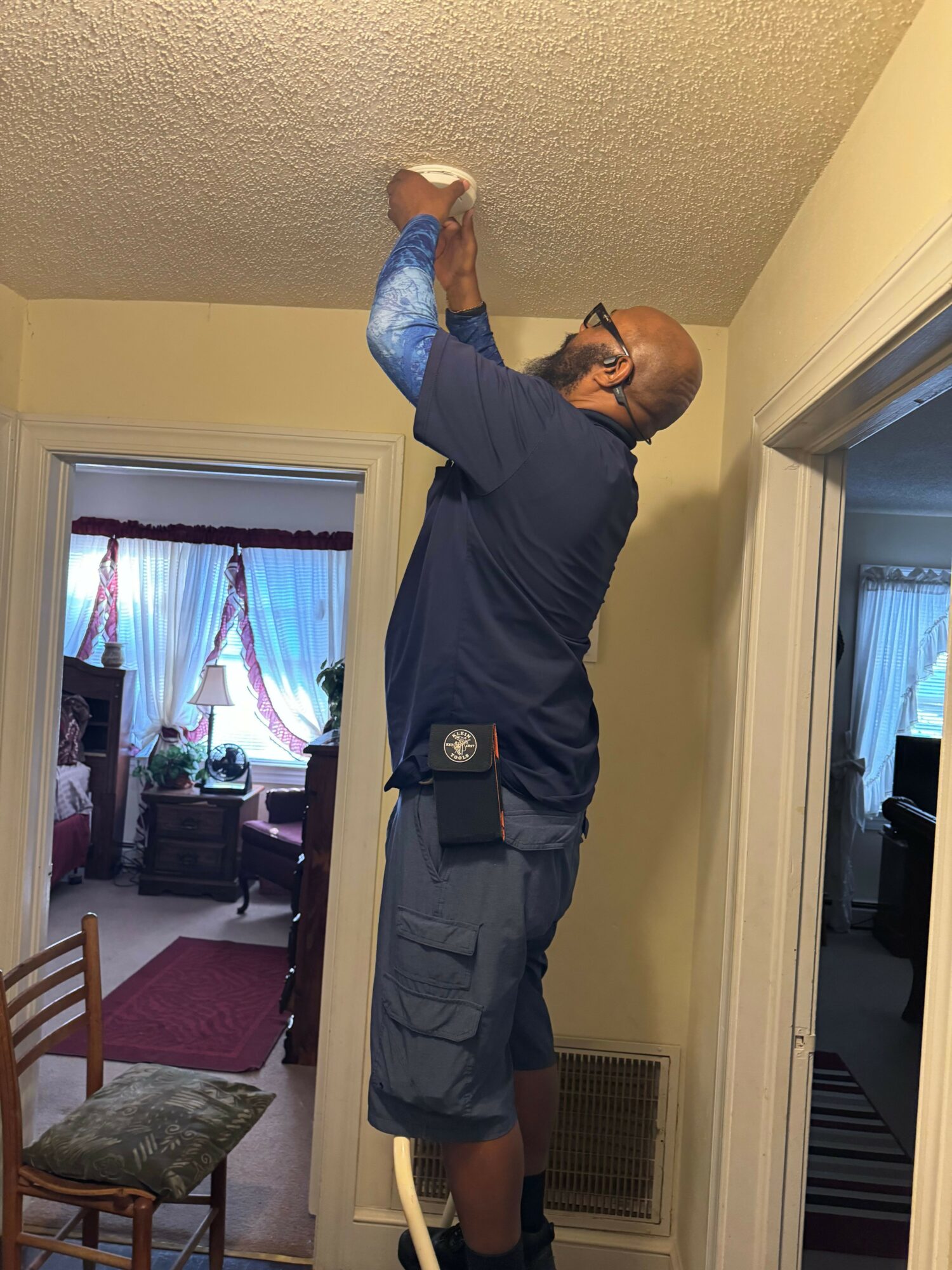 Man cleaning ceiling with cloth in living room, wearing glasses, dark shirt, and shorts.