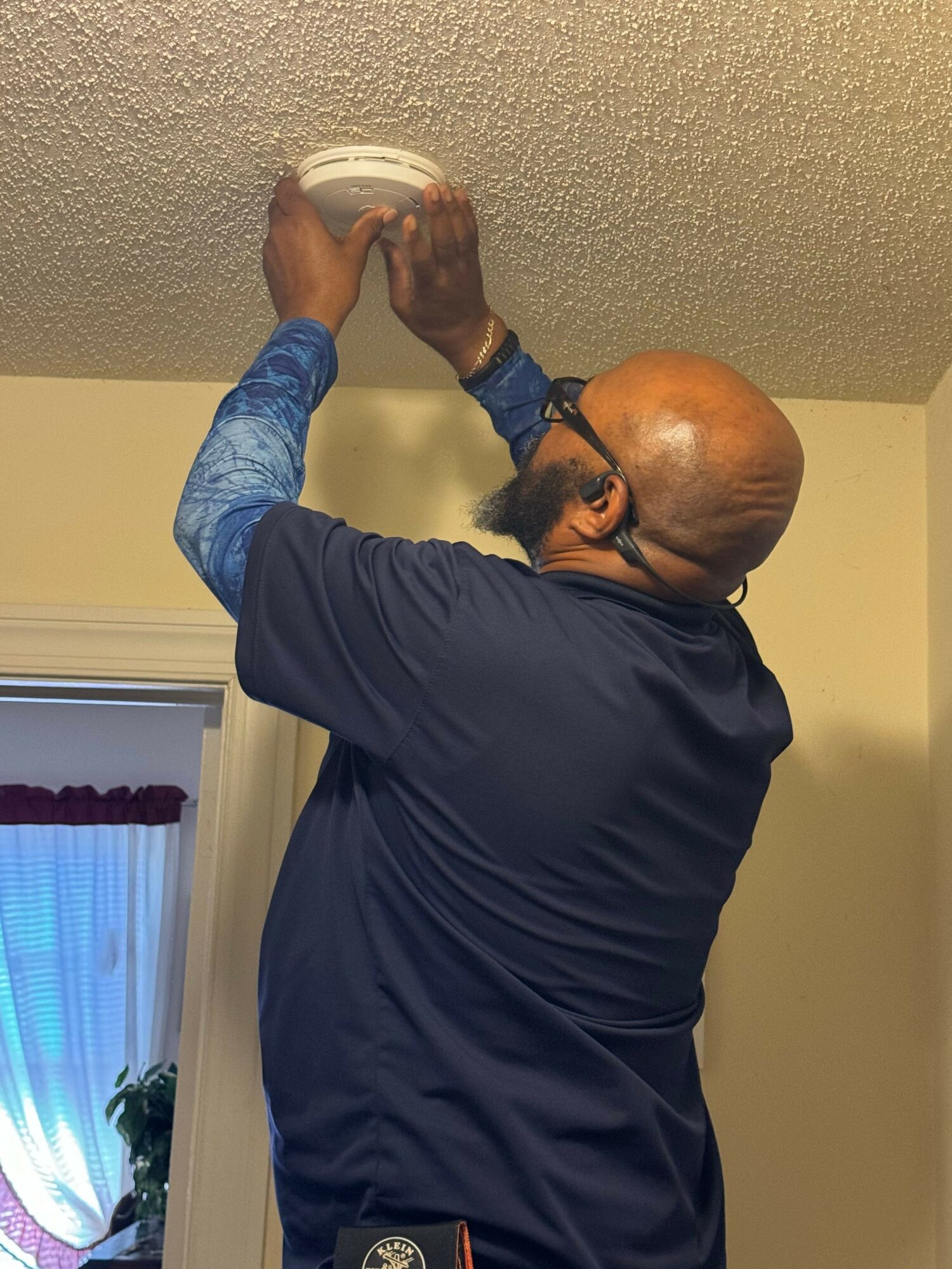 Person cleaning a textured ceiling with a cloth, wearing a dark shirt and blue sleeves, in a room with beige walls.