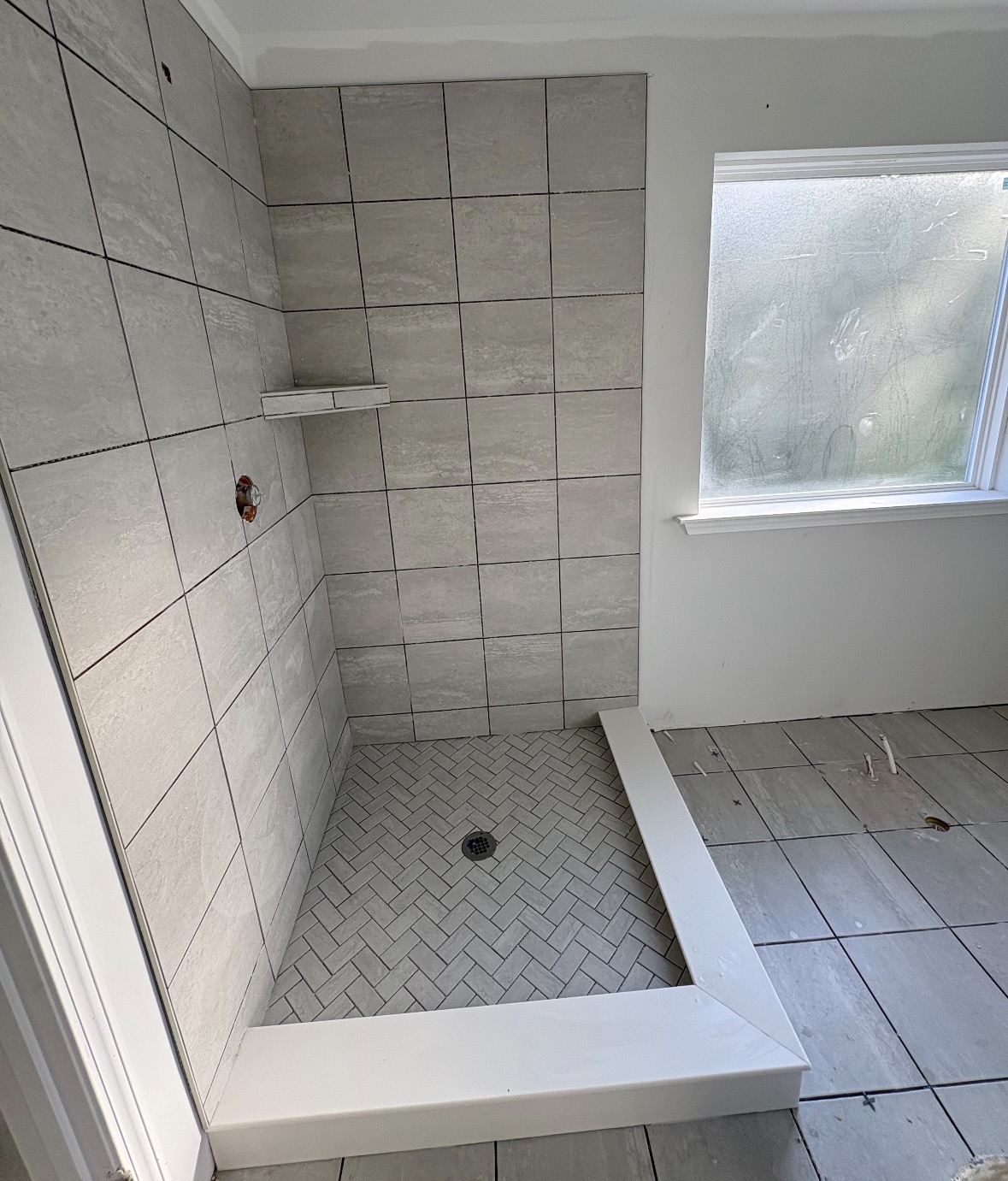 Shower area with tiled walls and floor, a small shelf, and a window nearby, with a drain in the floor.