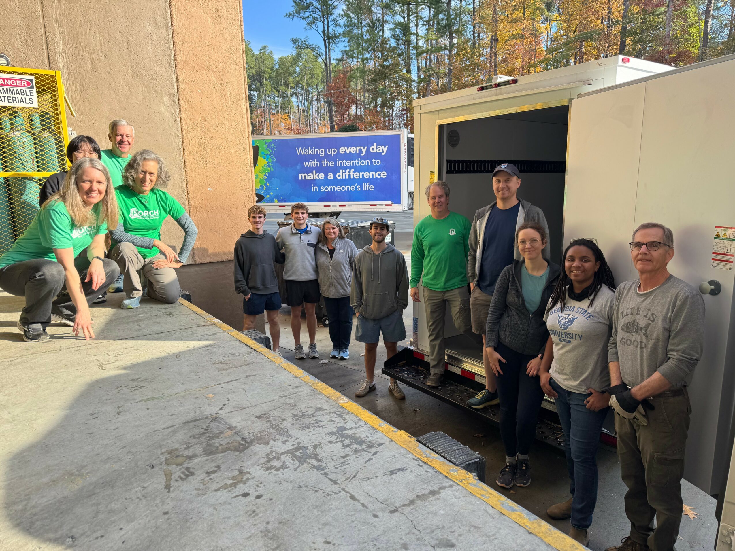 Group of people standing outside near a truck, some wearing green shirts, with trees and a building in the background.