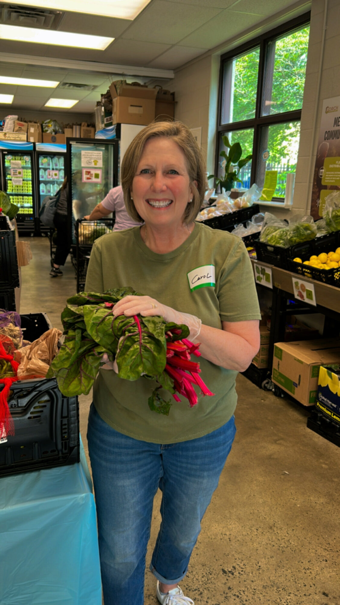 Smiling woman holding fresh vegetables in a grocery store, shelves and windows in background.