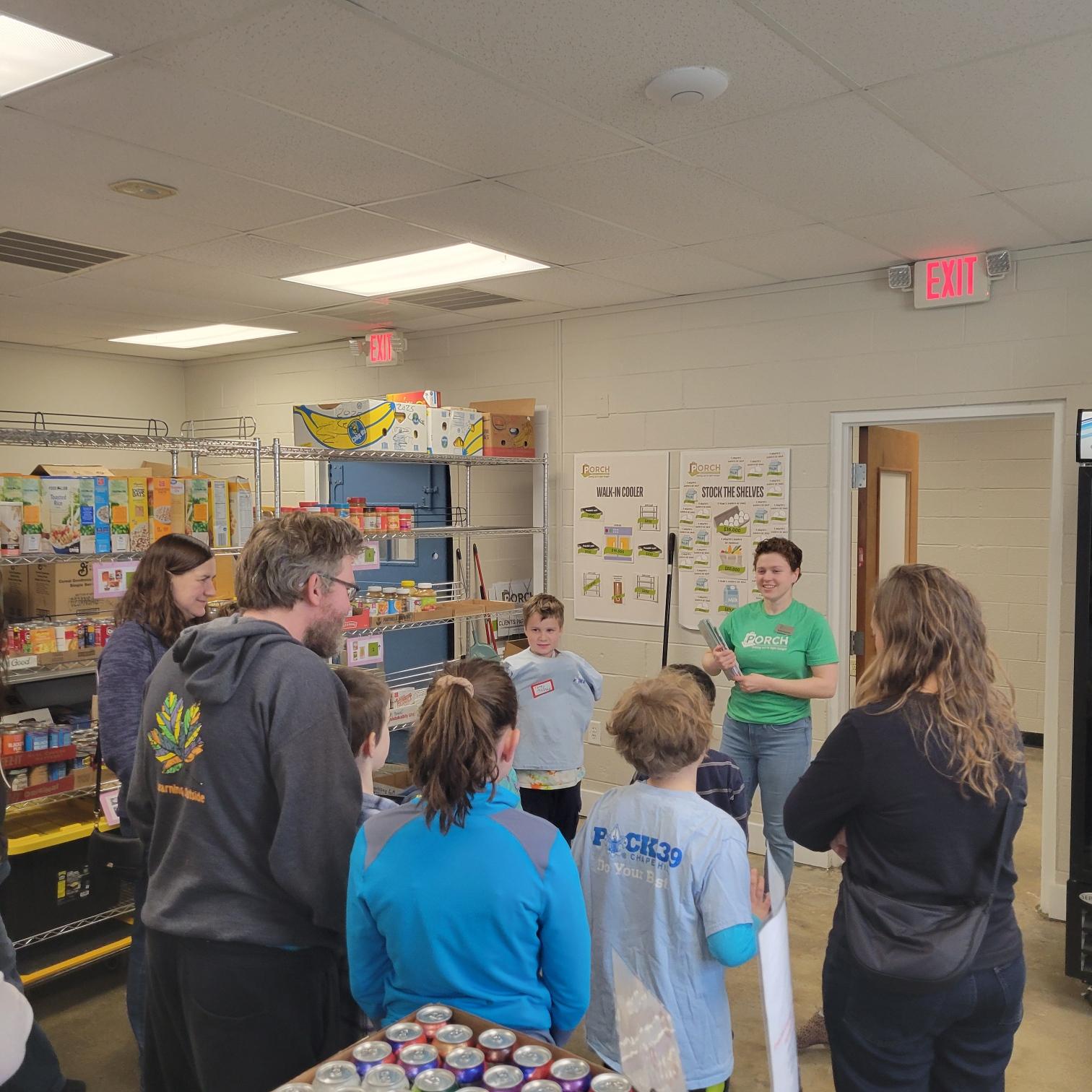 Group of children and a woman standing in a classroom, engaging in a discussion or activity, with shelves and posters in the background.