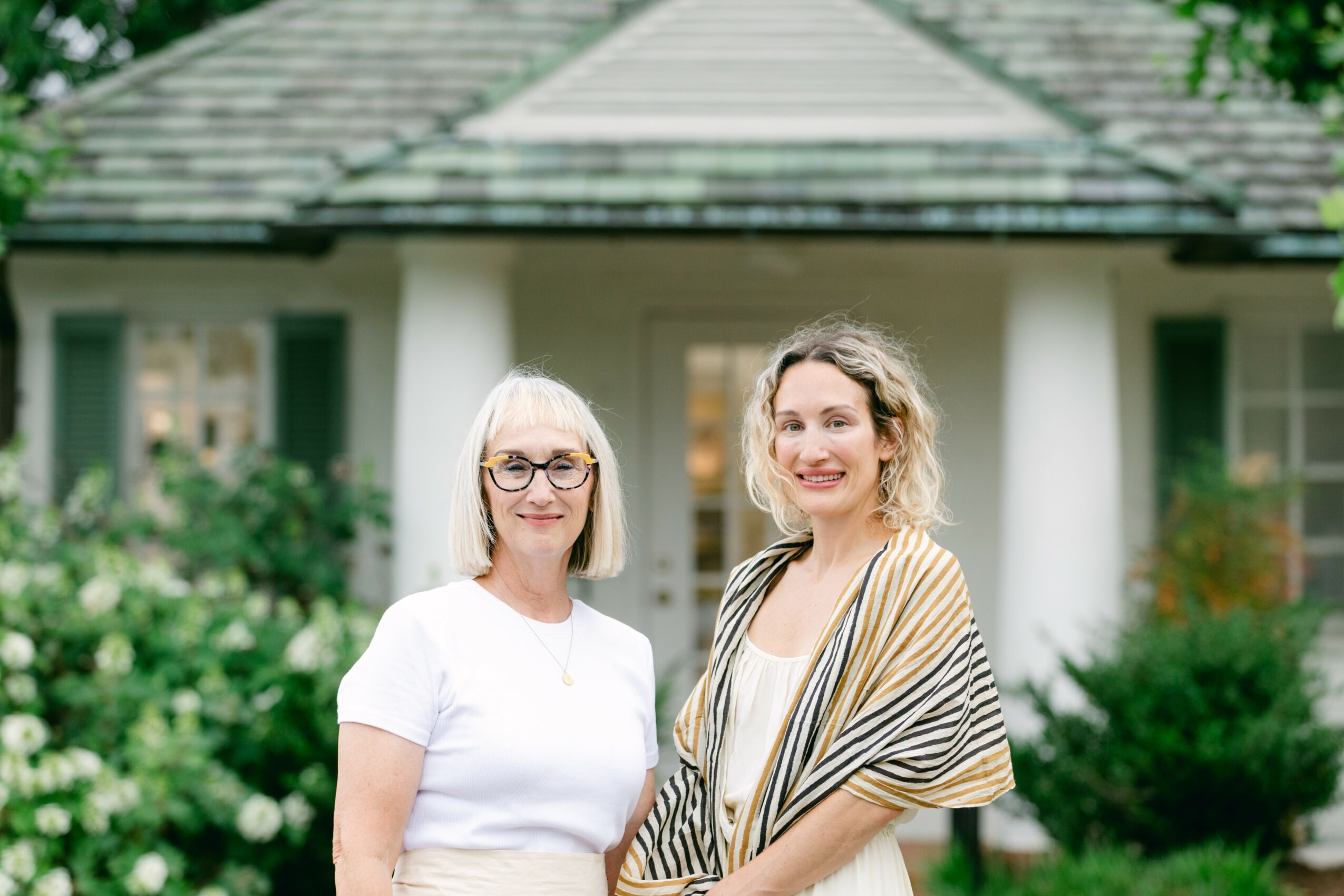 Two women standing outdoors in front of a house with white columns and greenery, smiling.