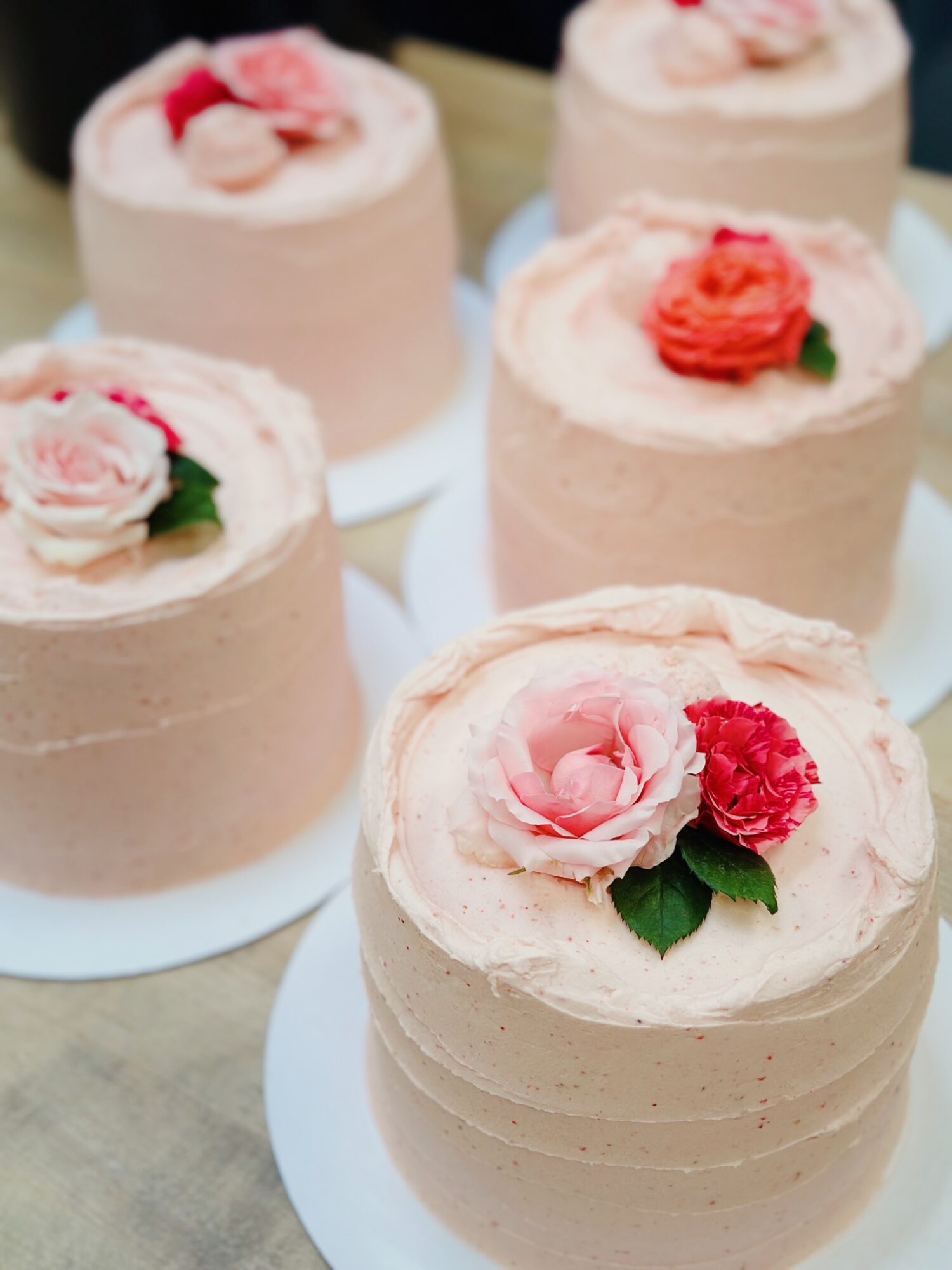 Six round cakes decorated with pink flowers and green leaves, on white bases, arranged on a wooden surface.