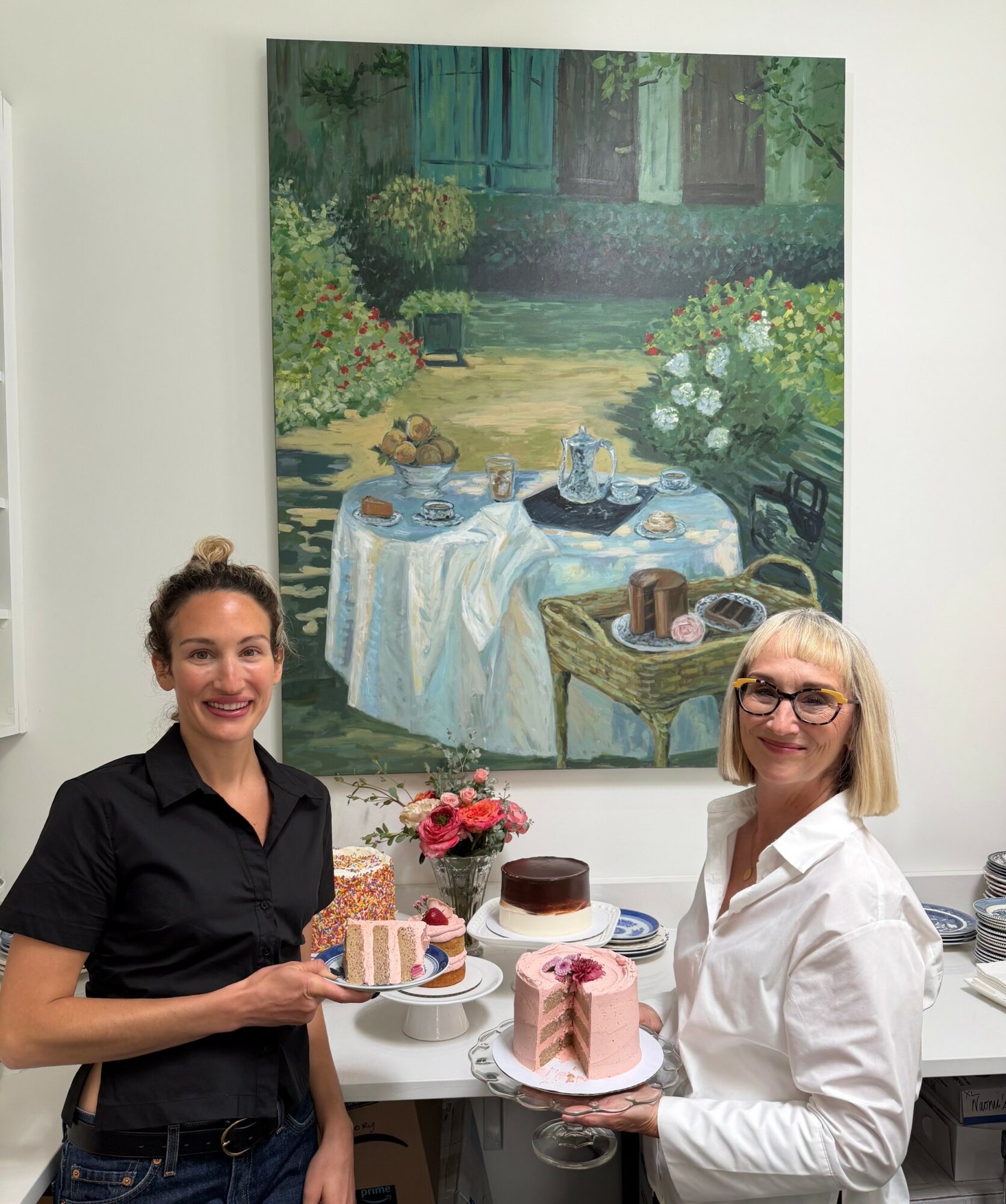 Two women stand in front of a table with cakes, in a room with a large outdoor scene painting on the wall.