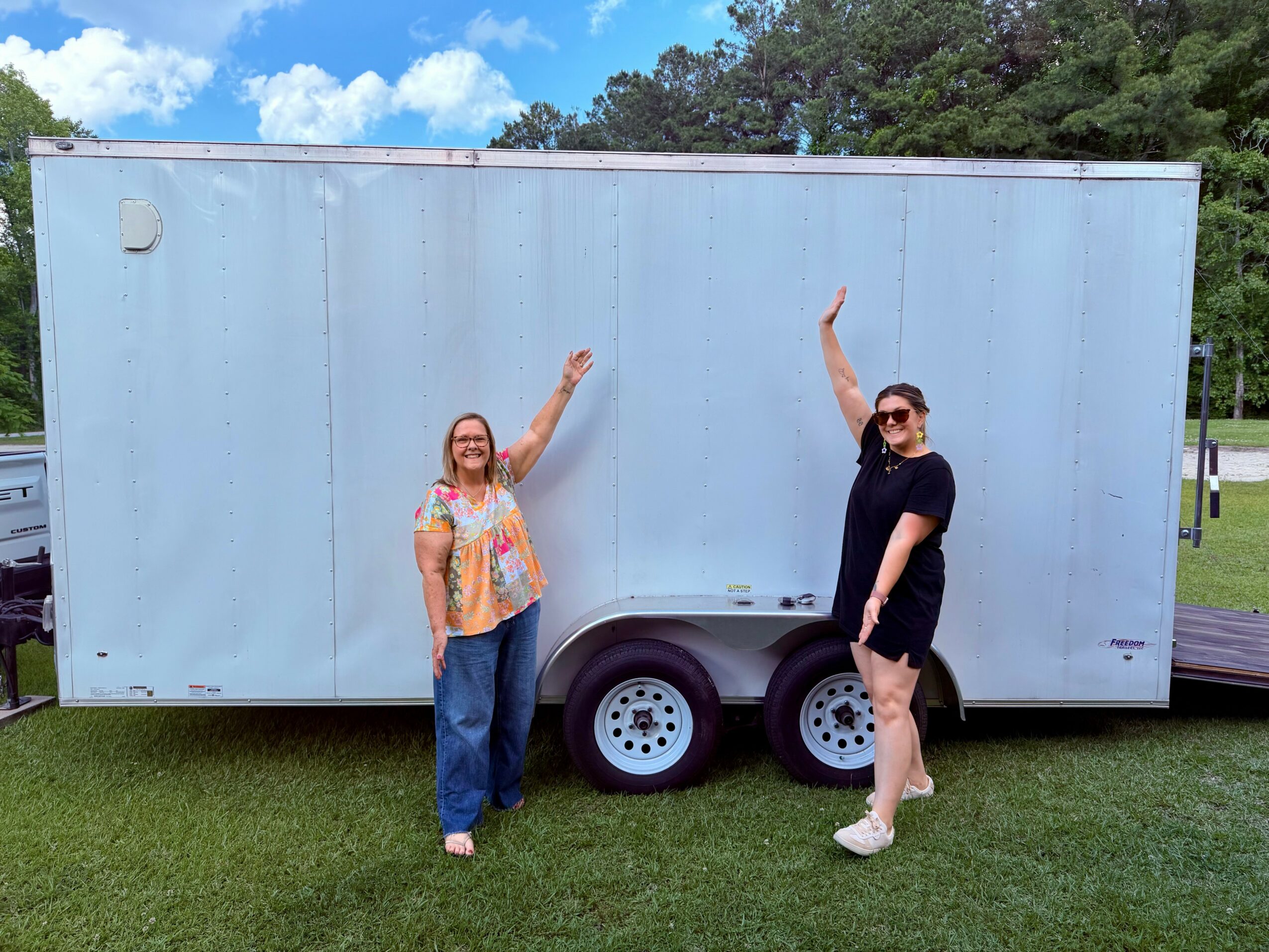 Two women stand in front of a large white trailer, waving and smiling outdoors on a grassy area with trees and blue sky.