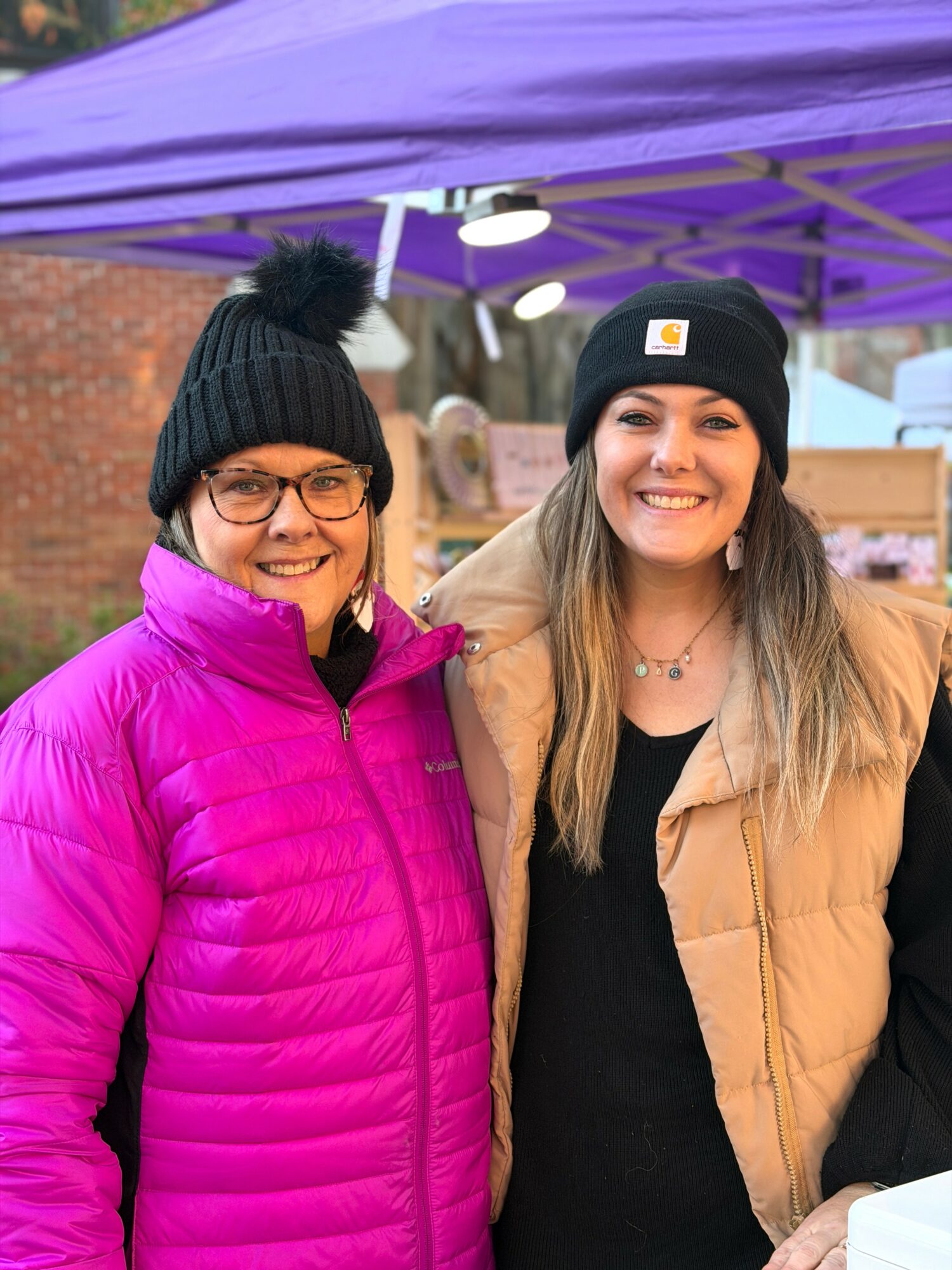 Two smiling women wearing winter hats and jackets outdoors, standing under a purple canopy.