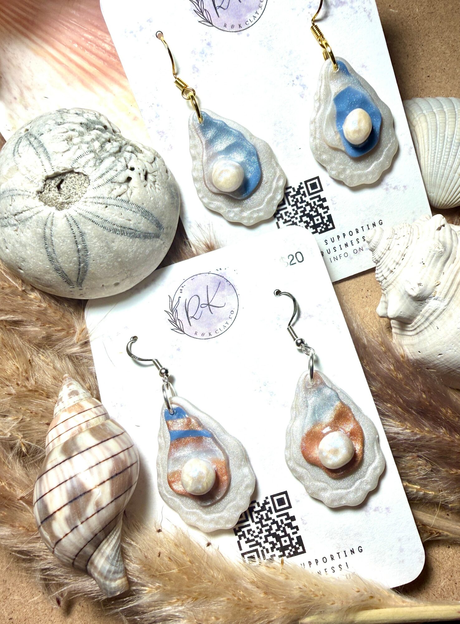 Two pairs of shell earrings with pearls, displayed on white cards with QR codes, surrounded by seashells and a stone.
