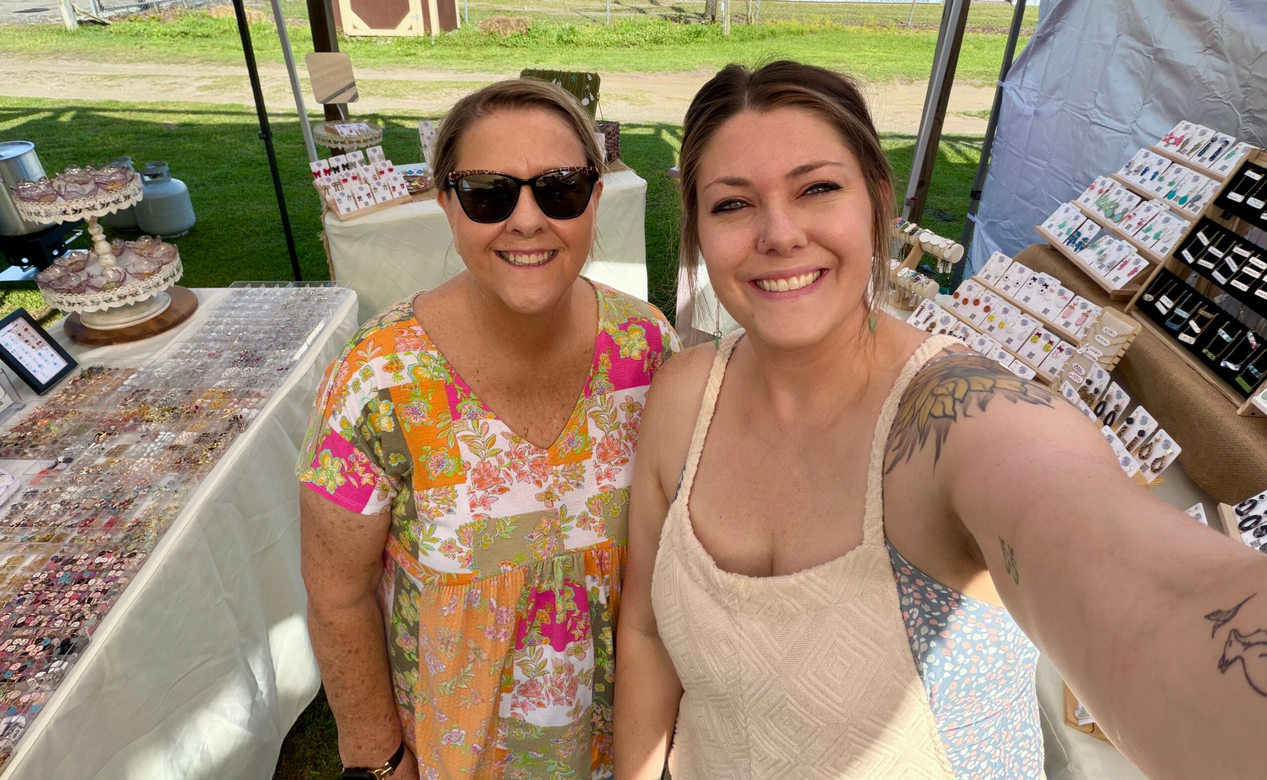 Two women smiling outdoors at a market stall with jewelry and crafts, one wearing sunglasses and a colorful dress, the other in a beige top.