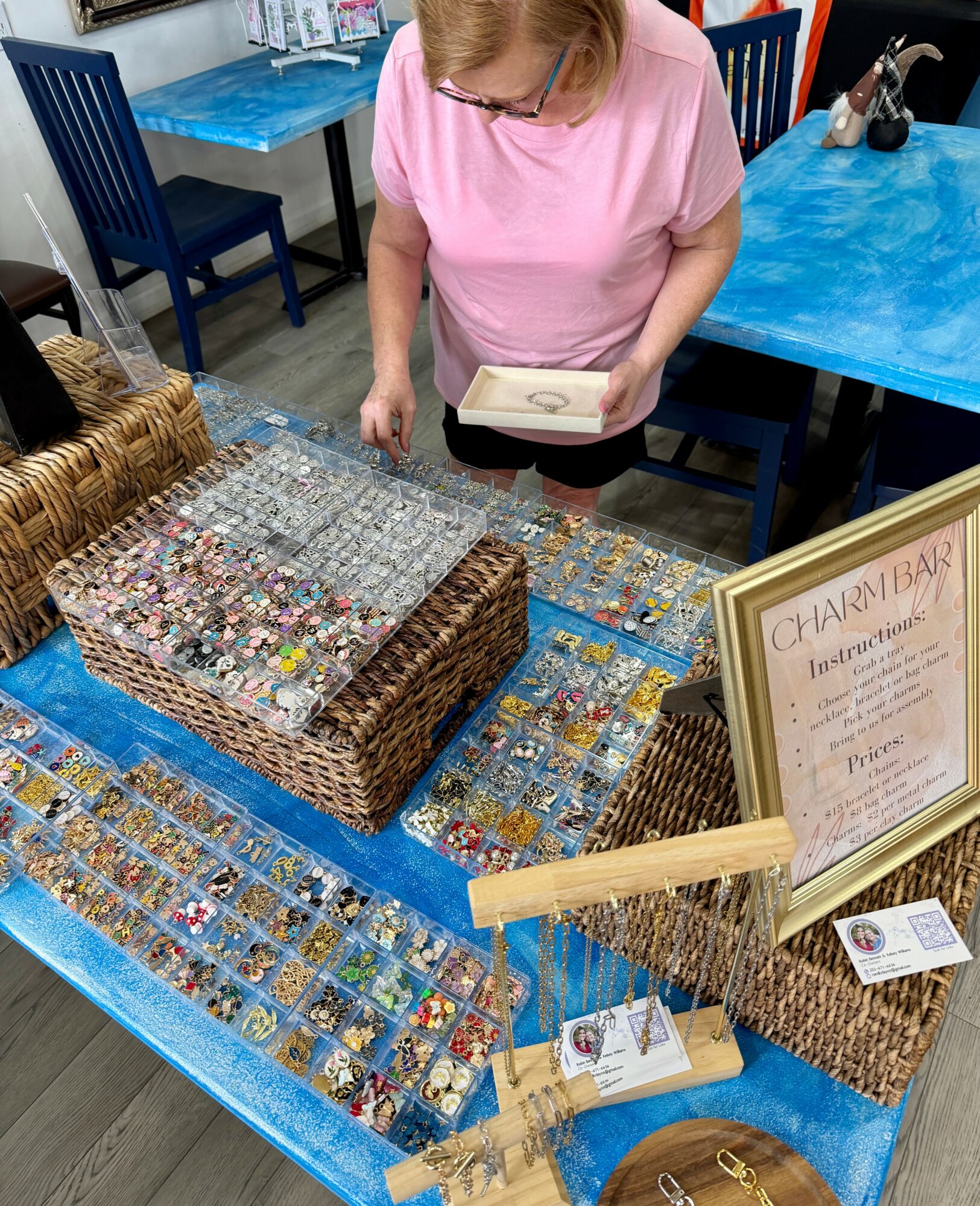 Woman in pink shirt looking at jewelry display on blue table, with baskets and a framed sign nearby.