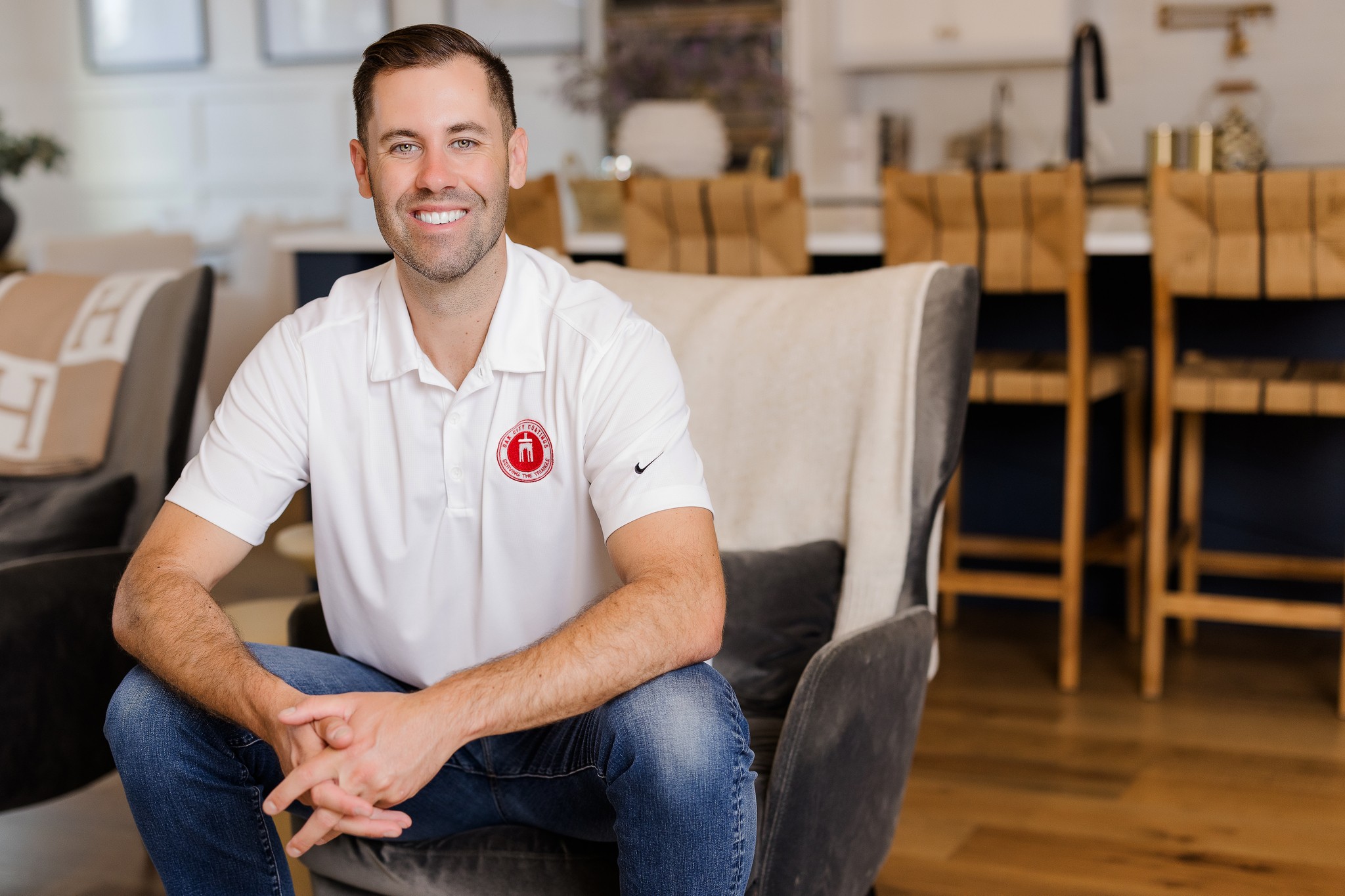 Smiling man sitting on a chair in a room with wooden floors and dining table in background.