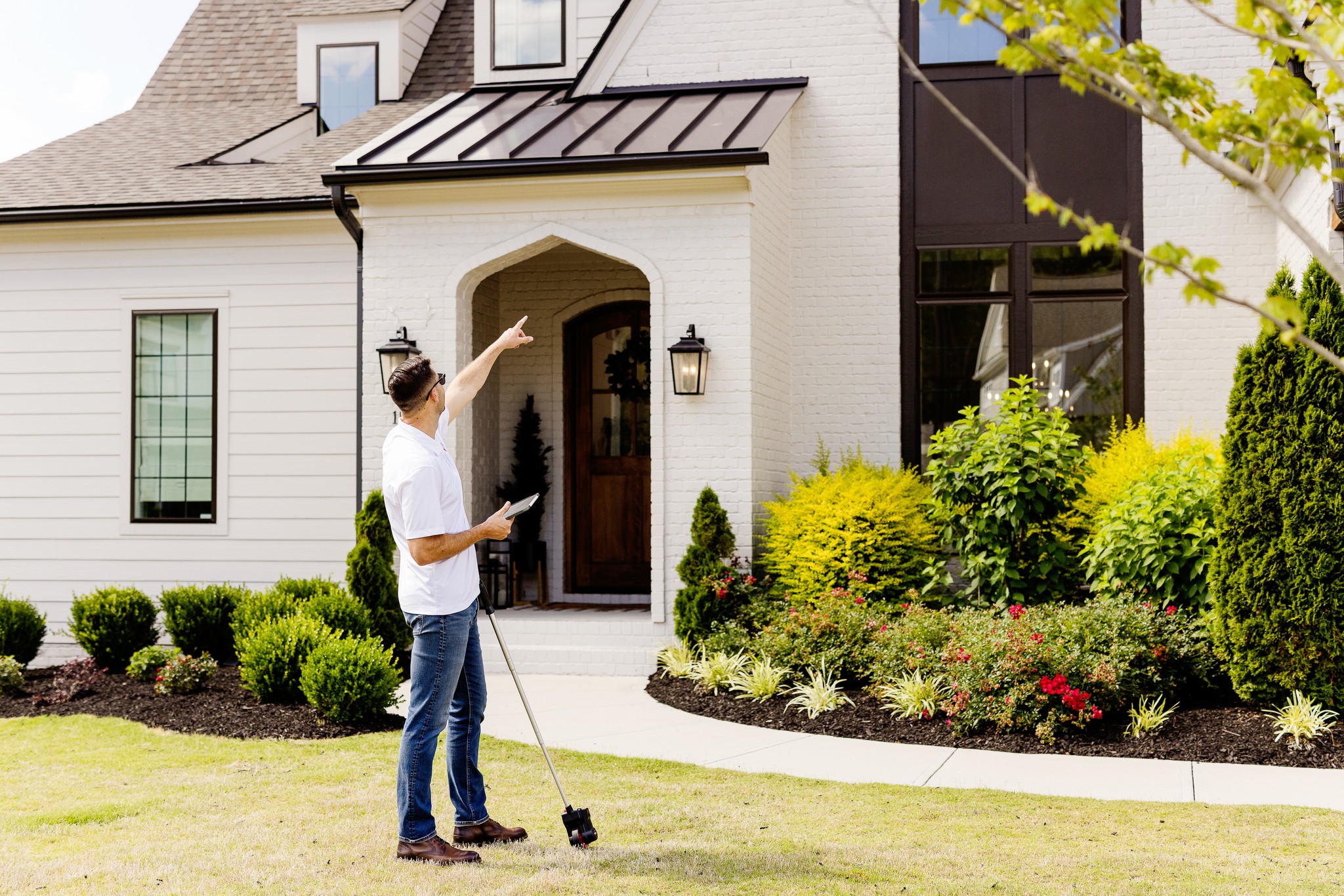 Person standing outside a house, pointing at the door, holding a remote, surrounded by landscaped garden.