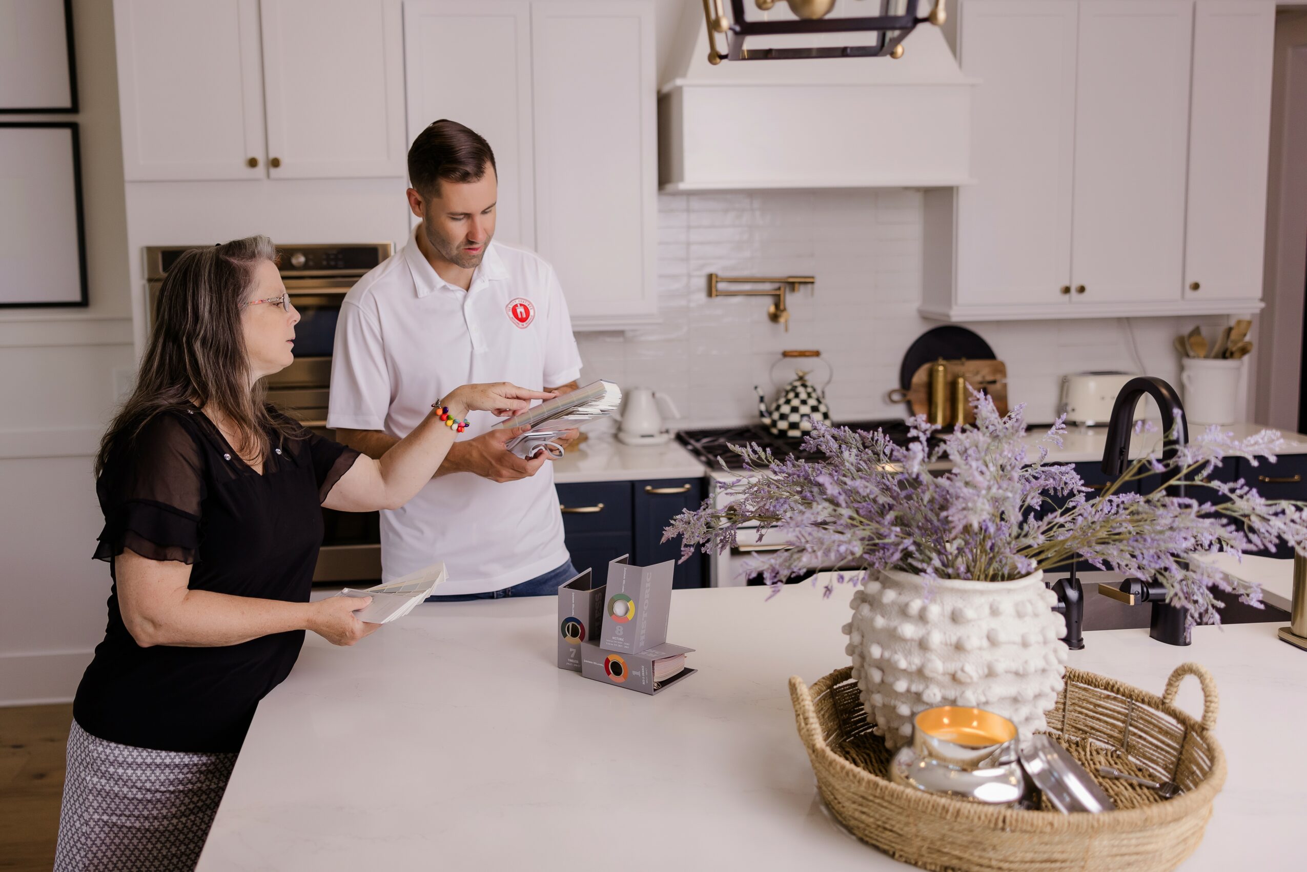 Two people stand at a kitchen counter, looking at a tablet and a magazine, with white cabinets and a large vase of flowers nearby.