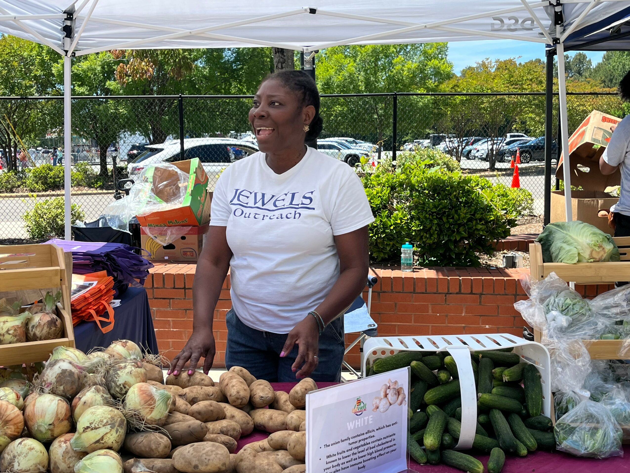 Smiling woman standing behind a table with vegetables at an outdoor market stall, with a tent overhead and cars in background.