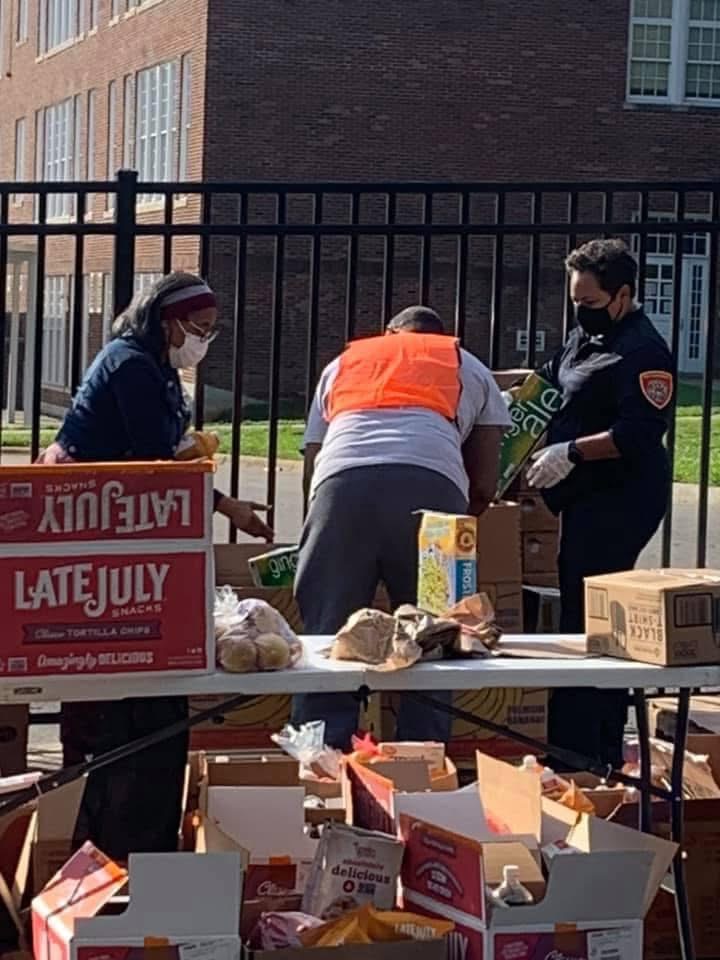 Three people, two women and one man, are working together at an outdoor table with boxes and food items, wearing masks.