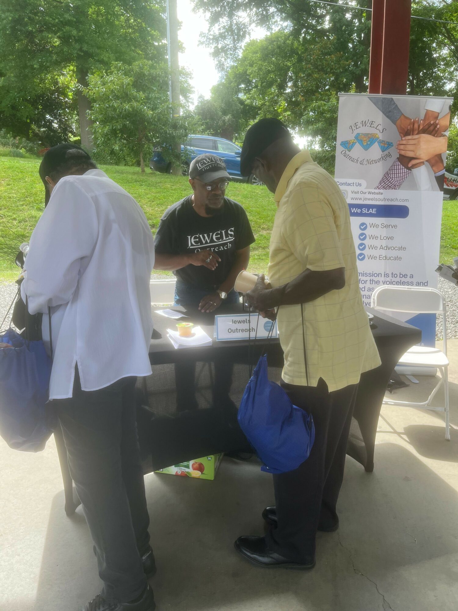 Two people standing outdoors near a table with informational materials, trees and a banner in the background.