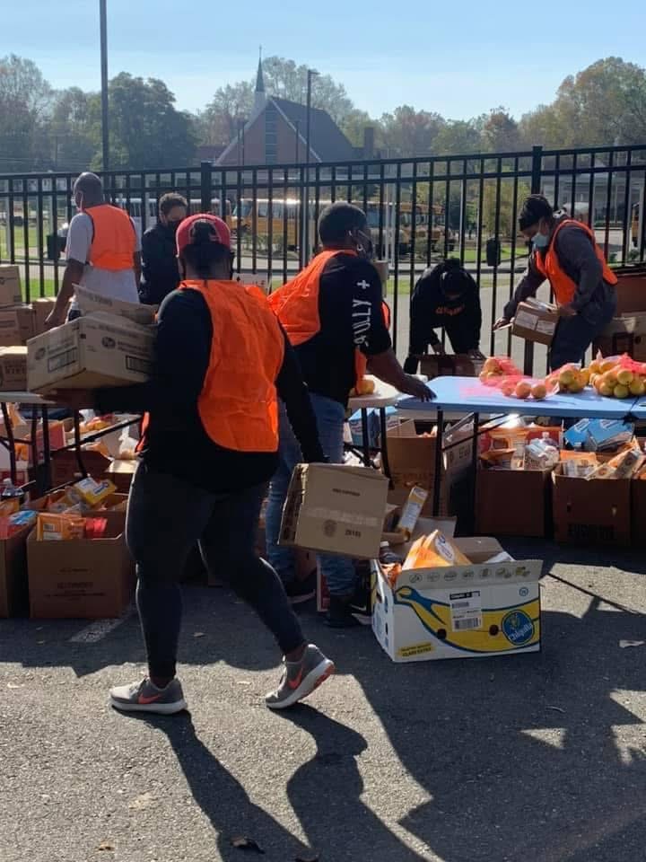 People in orange vests unload boxes of supplies outdoors on a sunny day, with a fence and trees in the background.