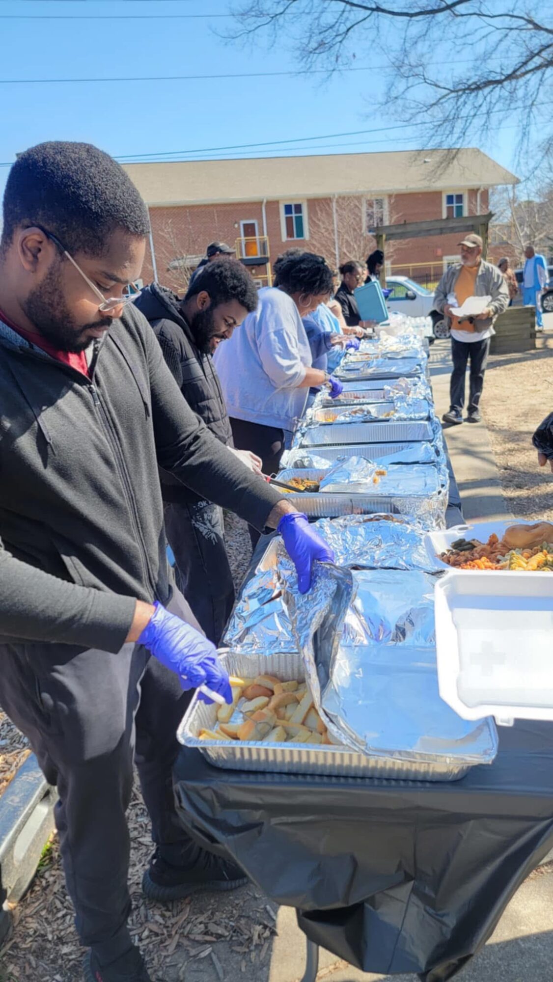 People line up outdoors at a table with food trays, serving food on a sunny day, with a building and trees in background.