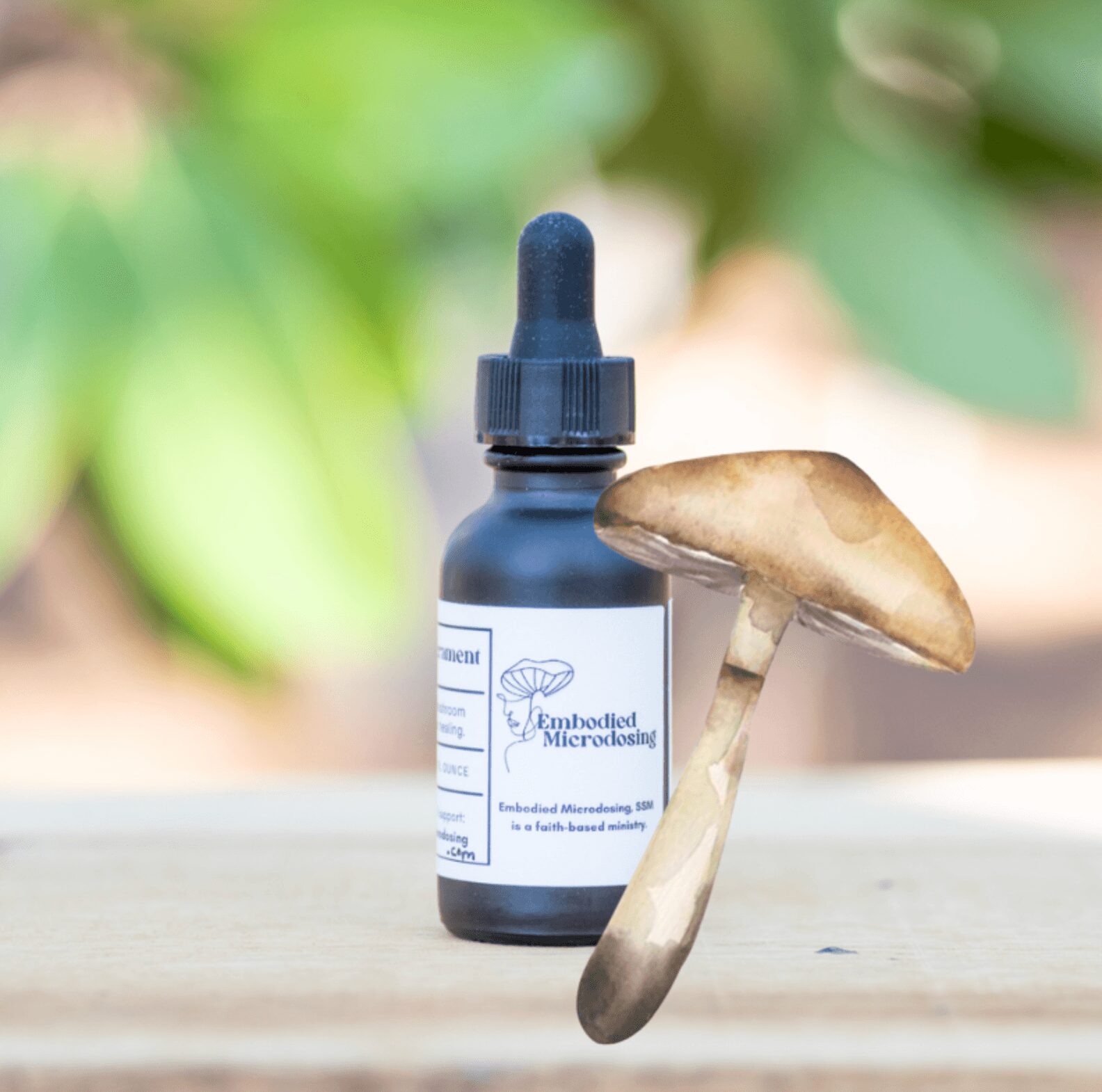 Dropper bottle labeled Embodied Microdosing next to a mushroom on a wooden surface, with blurred green foliage background.