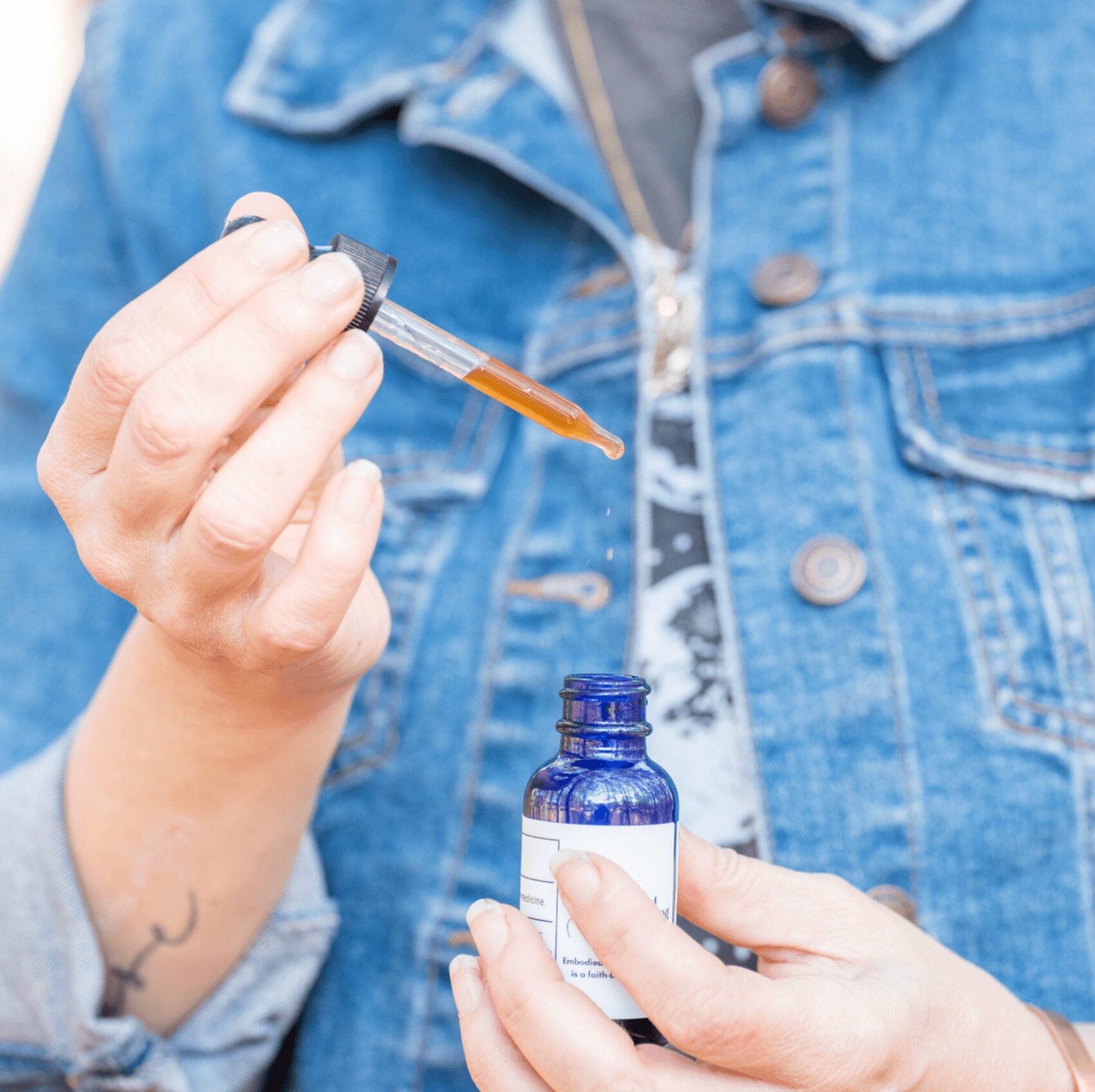 Person in denim jacket holding a blue bottle and using a dropper with brown liquid.