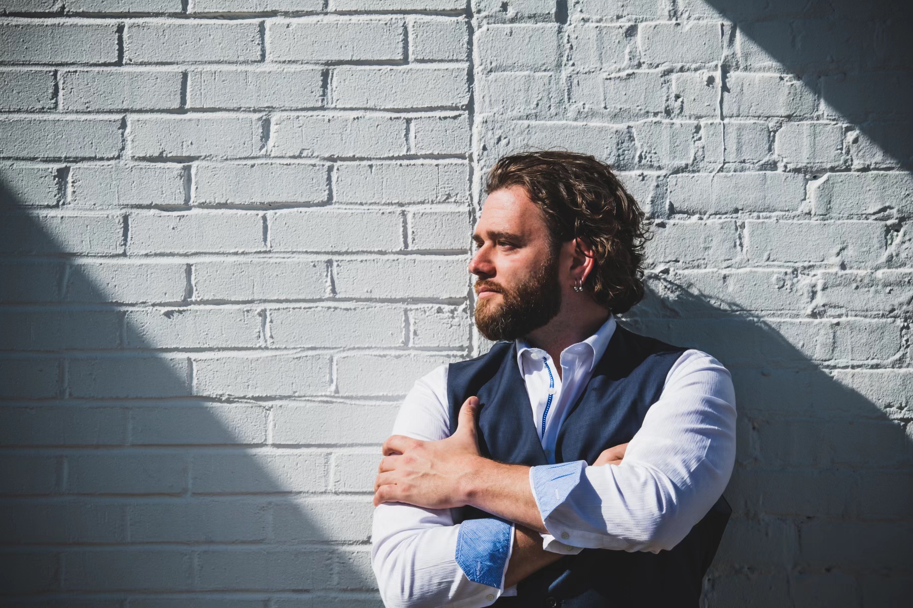 Man with beard and curly hair leaning against a white brick wall, arms crossed, looking to the side, sunlight casting shadows.