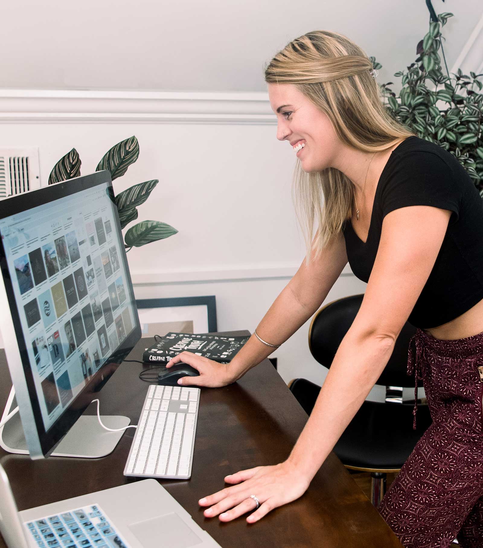 Woman smiling at computer screen in a bright room with plants, leaning forward with one hand on the desk.