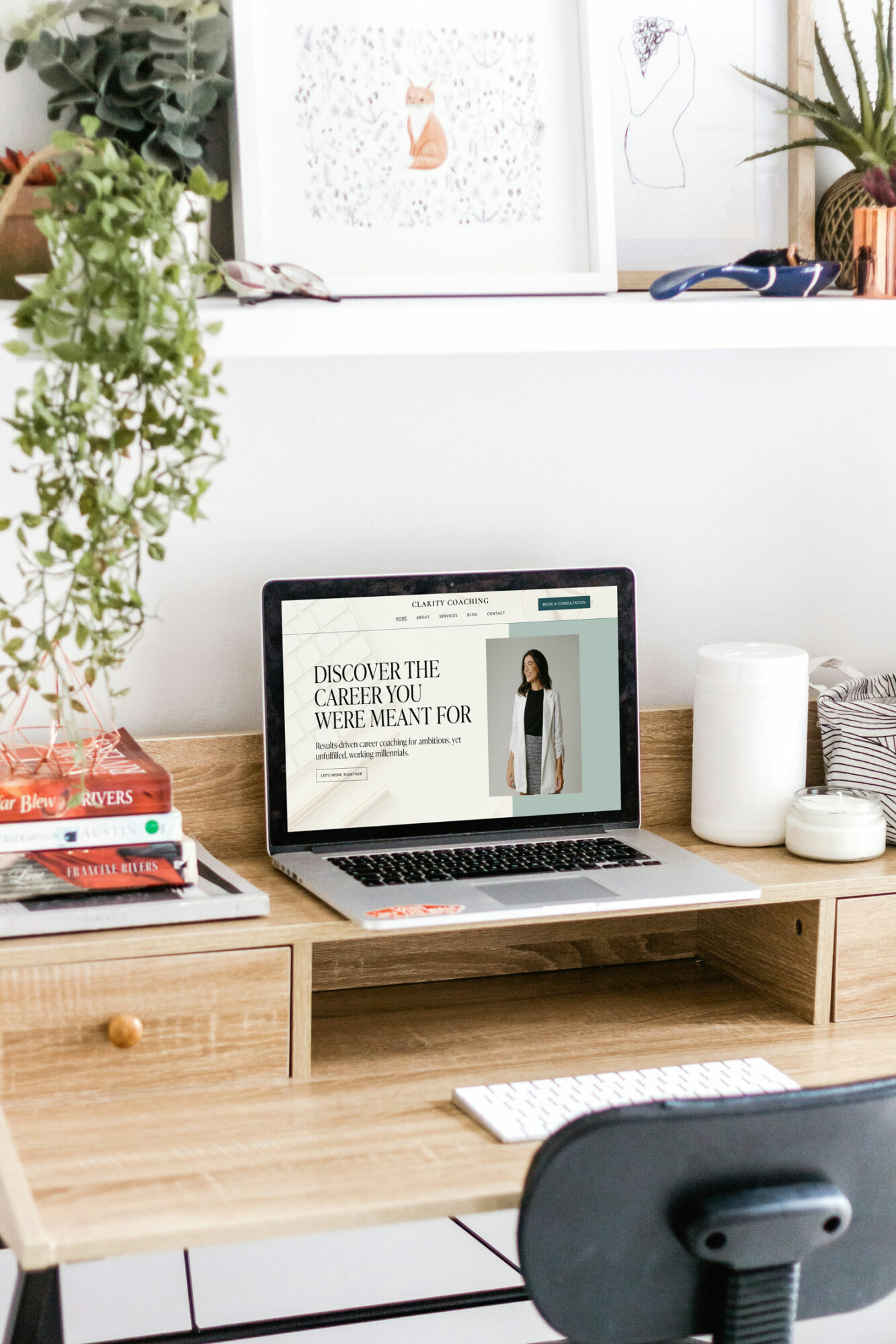 Laptop on a wooden desk displaying a website, with books, plants, and decorative items in a bright room.