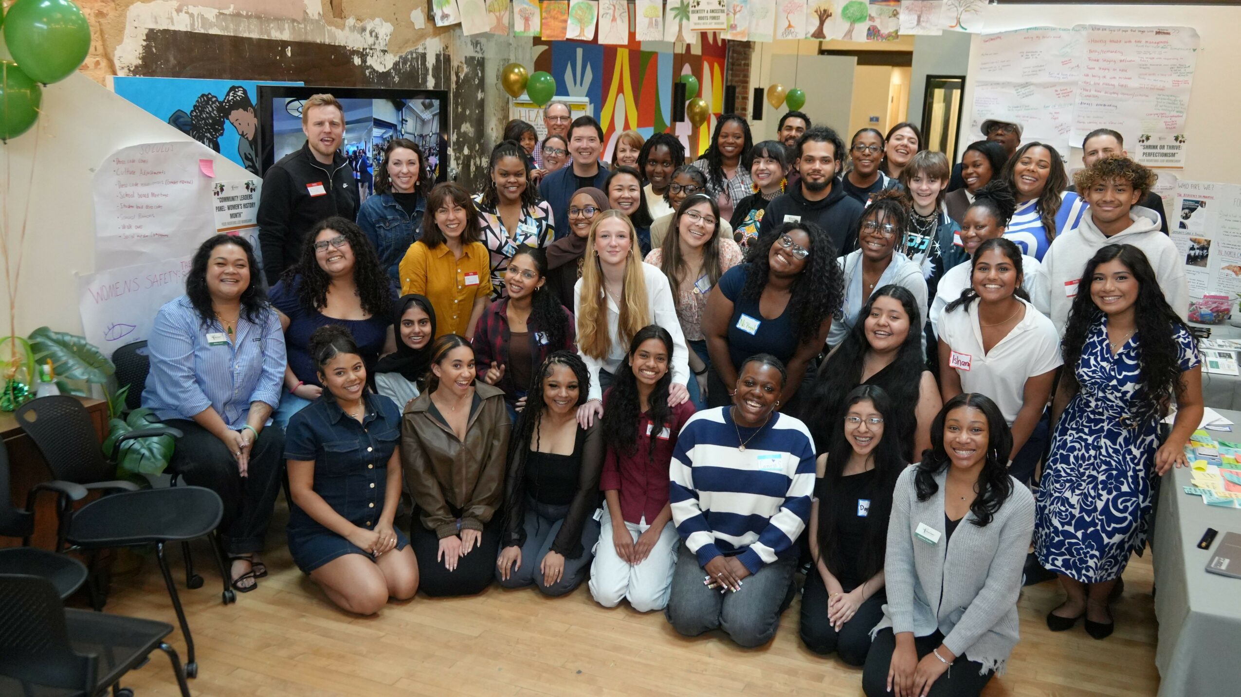 Group of diverse people posing together in a room with colorful decorations and a whiteboard, smiling at the camera.