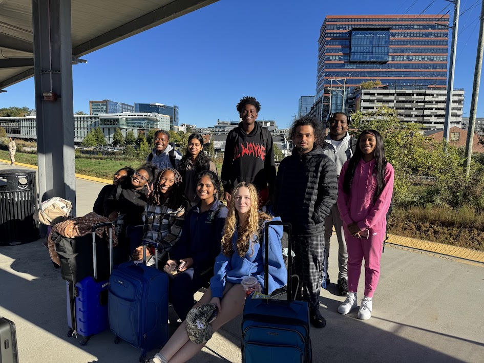 Group of ten people standing outdoors with luggage, city buildings in background, sunny day.
