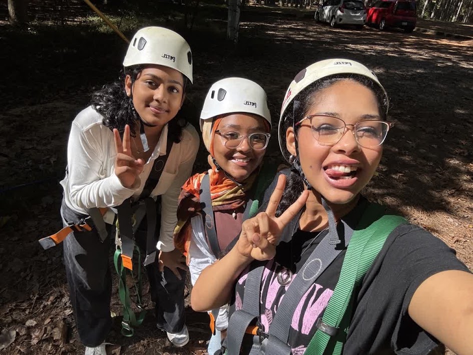 Three children wearing helmets and harnesses outdoors, smiling and making peace signs, with trees and parked cars in background.