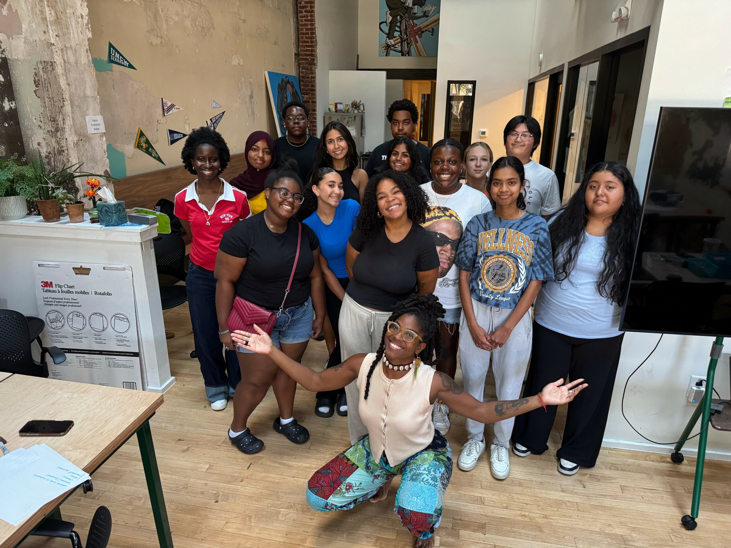 Group of 14 diverse women and girls standing together indoors, smiling, some with arms around each other, in a casual setting.