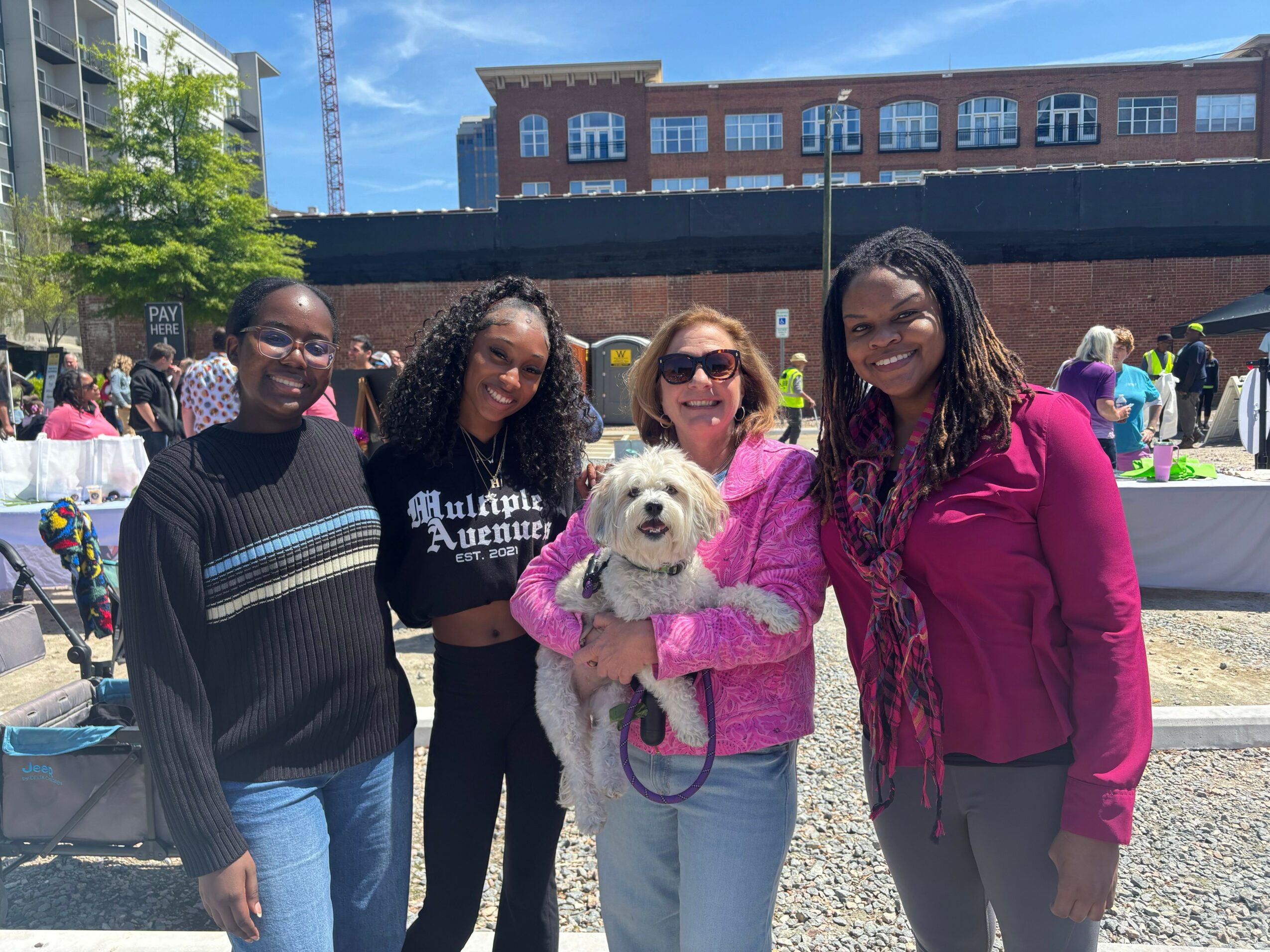 Three women and a small dog outdoors at a market or fair, with buildings and tents in the background.