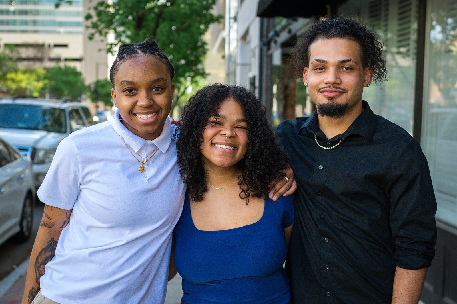 Three smiling people standing outdoors, two women and one man, with buildings and trees in the background.