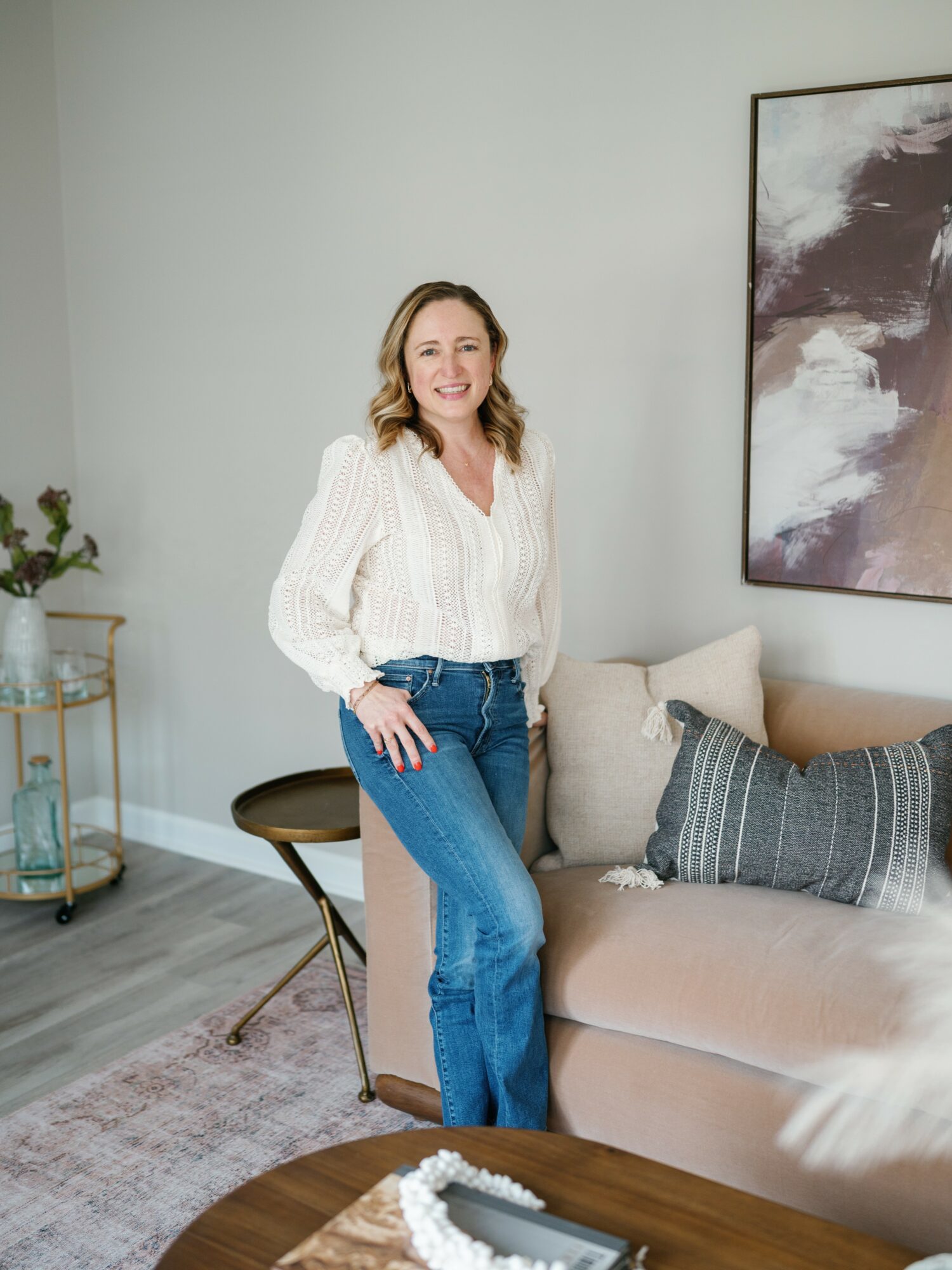 Woman with shoulder-length hair smiling, standing next to a beige sofa with pillows in a living room.