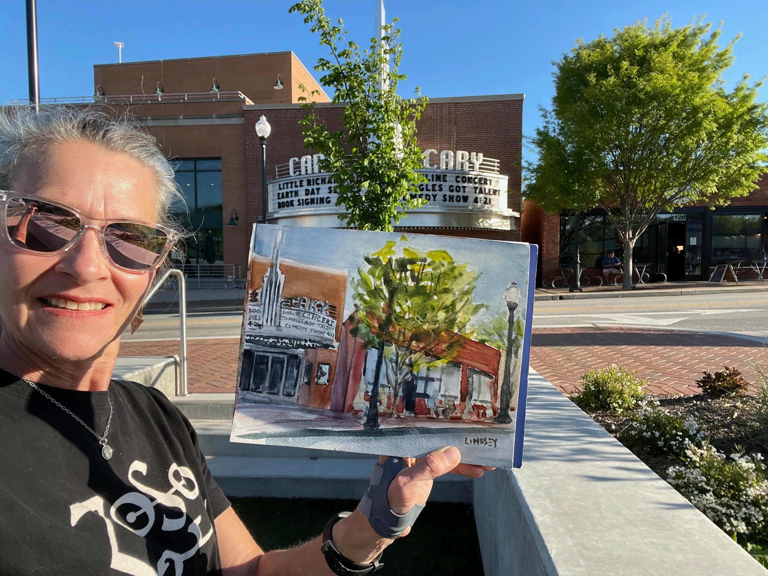 Woman with sunglasses holding a painting of a street scene with trees and buildings outdoors on a sunny day.